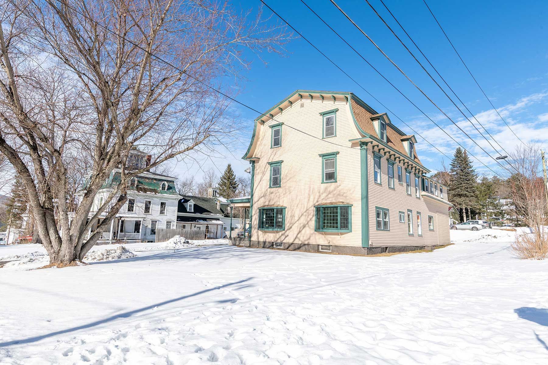 Snow-covered beige house with green-trimmed windows under a blue sky, beside bare trees and power lines