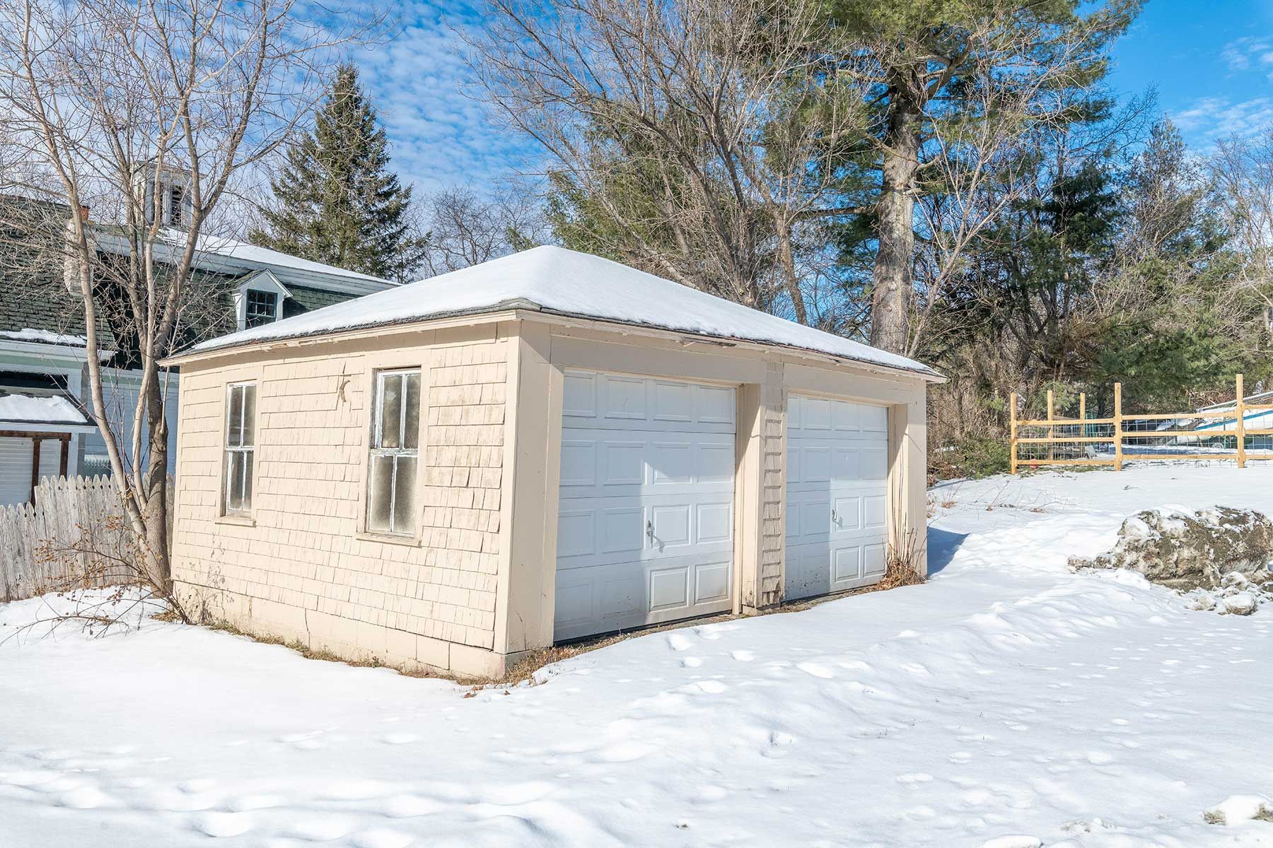 Snow-covered beige garage with two white doors in a wooded backyard setting