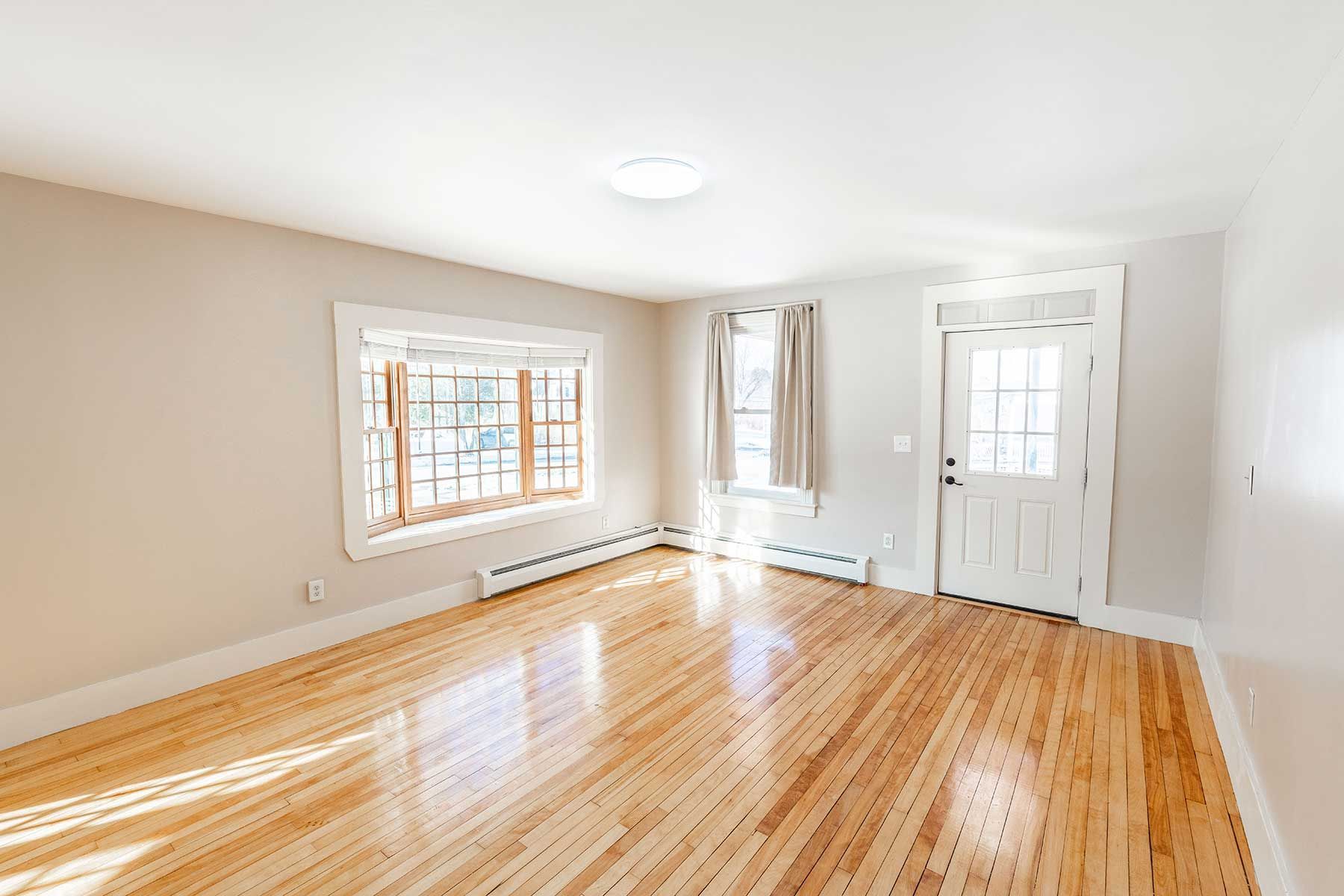 Empty bright rental room with hardwood floors, beige walls, window, and white front door