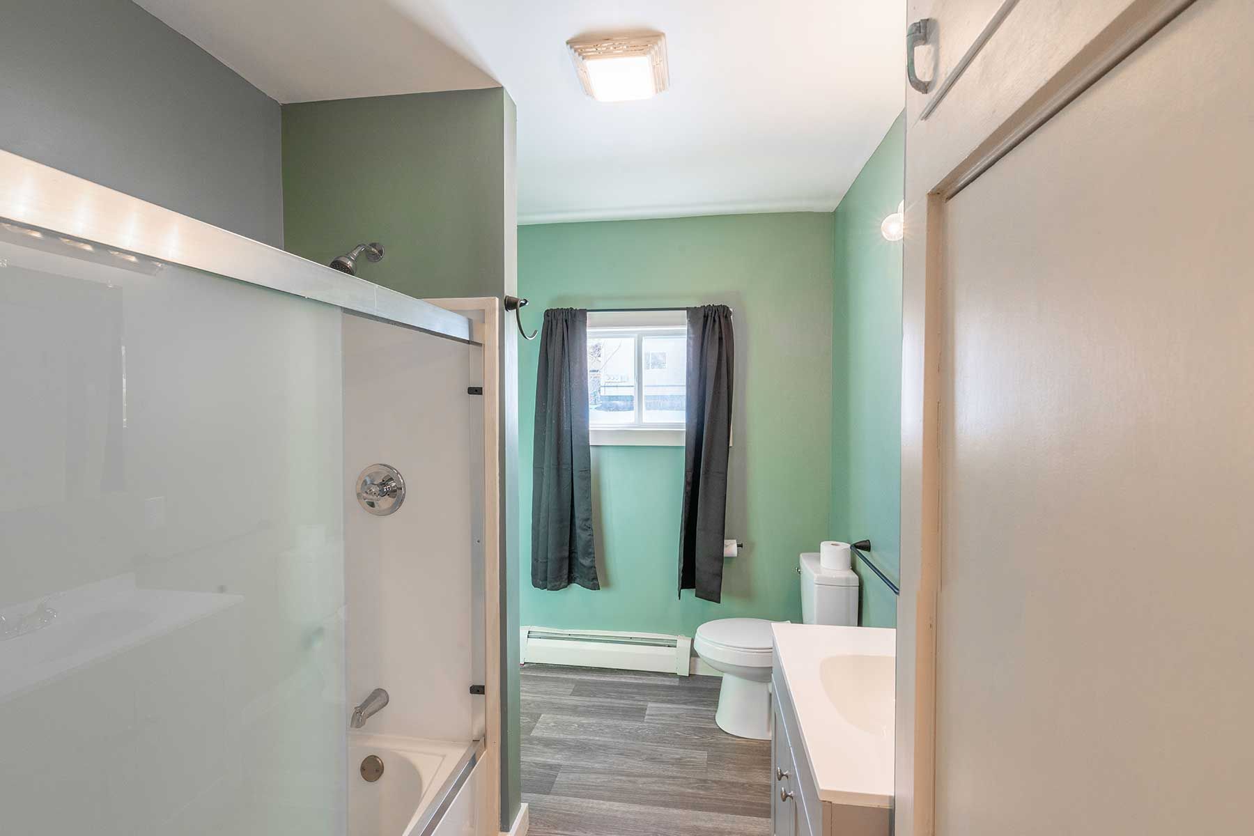 Narrow bathroom with white fixtures, mint green walls, and window with dark curtains