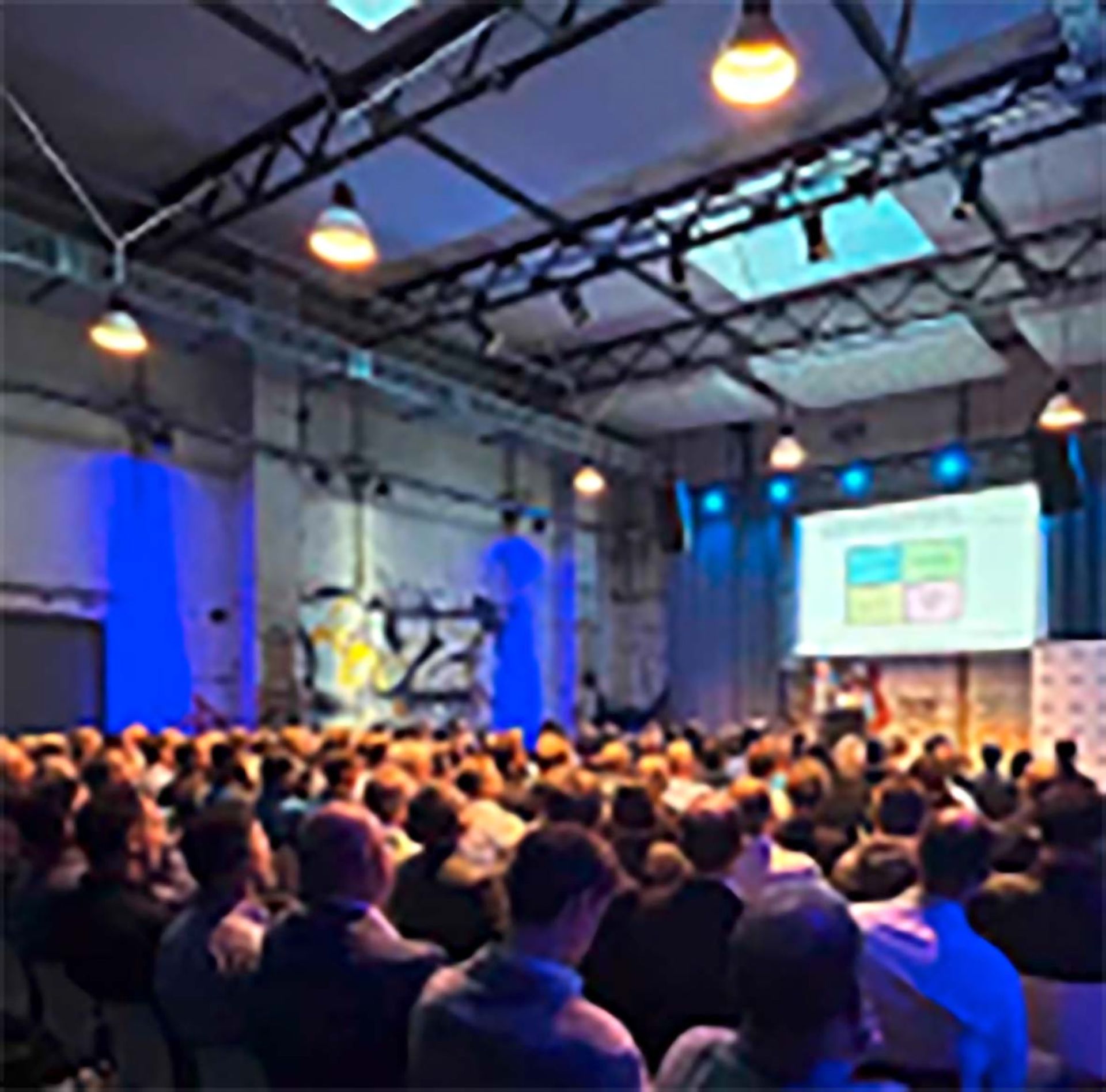 In einem modernen Saal im Industriestil mit blauer Akzentbeleuchtung sitzt ein großes Publikum vor einem Moderator und einer Projektionsfläche.
