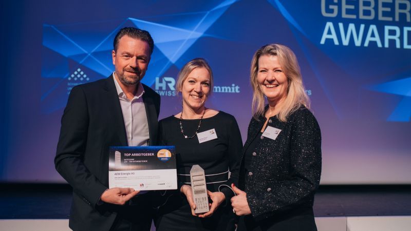 Drei Fachleute stehen lächelnd auf der Bühne bei einer Preisverleihung und halten einen Preis und eine Urkunde in den Händen.