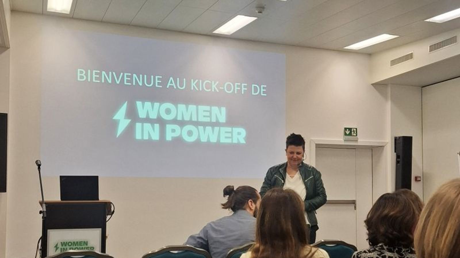 Eine Rednerin spricht zu einem Publikum in einem Raum, auf dem eine Leinwand mit der Aufschrift „BIENVENUE AU KICK OFF DE WOMEN IN POWER“ projiziert wird.