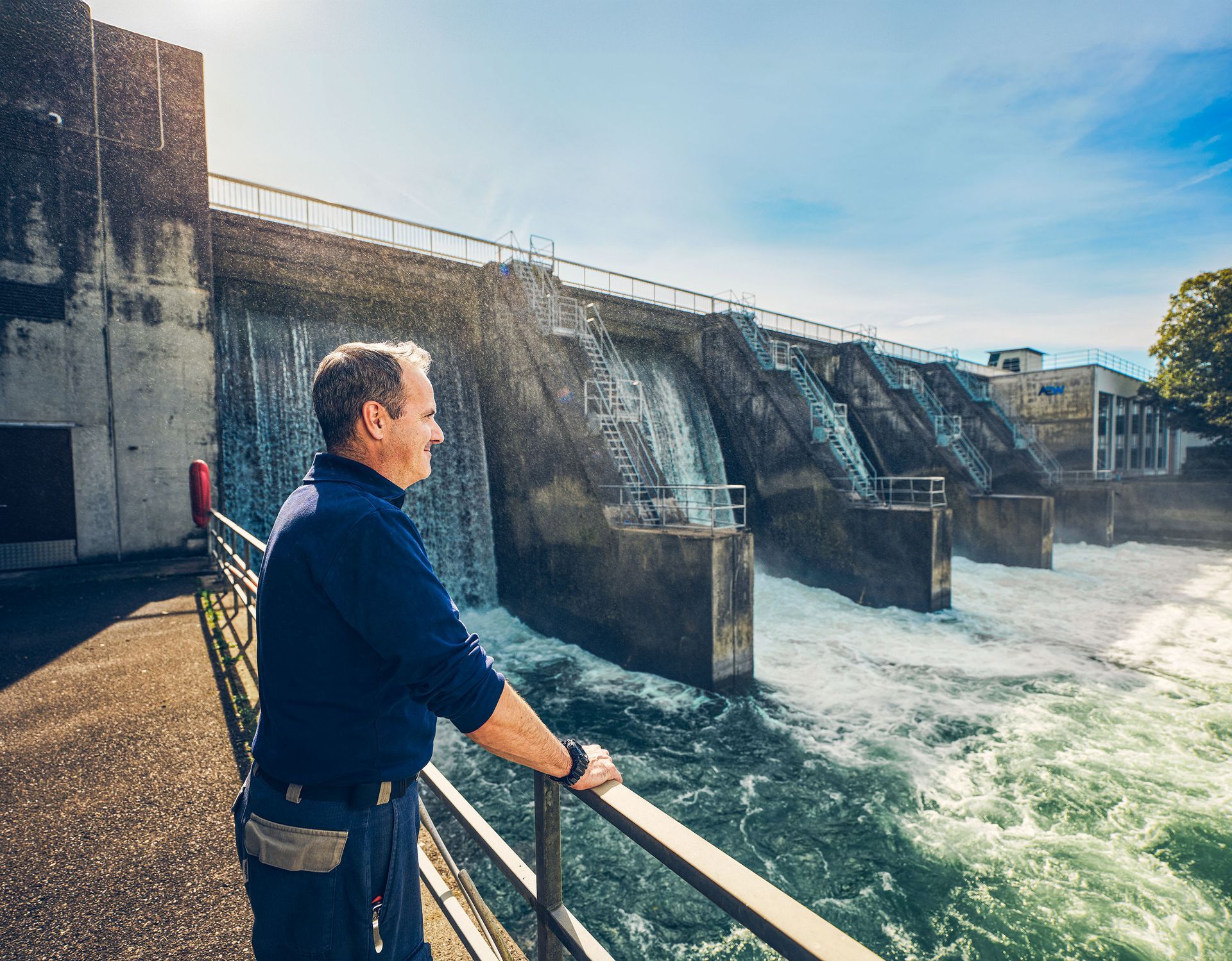 Eine Person in blauer Uniform steht an einem Metallgeländer und blickt auf einen Staudamm, über dessen mehrere Überlauftore Wasser fließt.