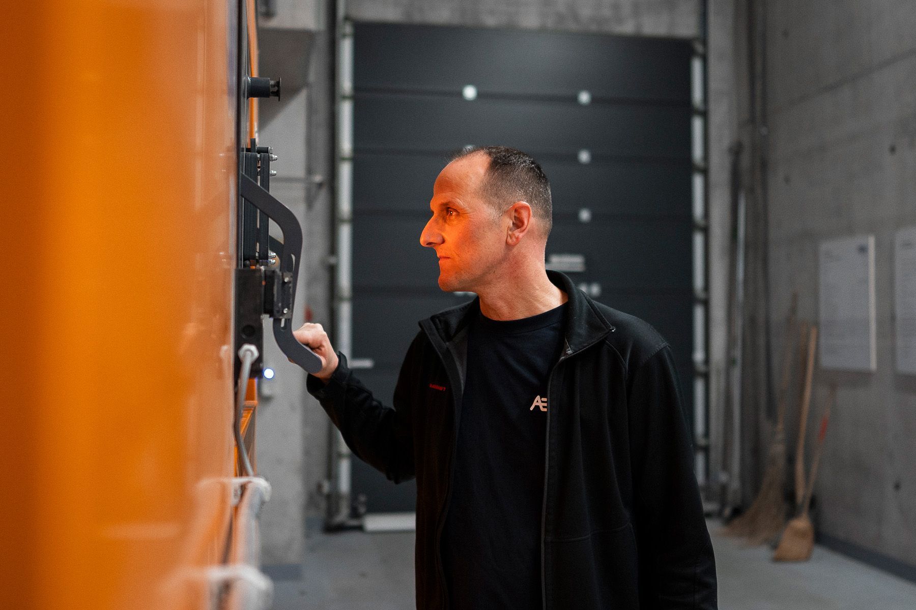 Eine Person in einer schwarzen Reißverschlussjacke inspiziert ein orangefarbenes industrielles Bedienfeld in einer Betonhalle.