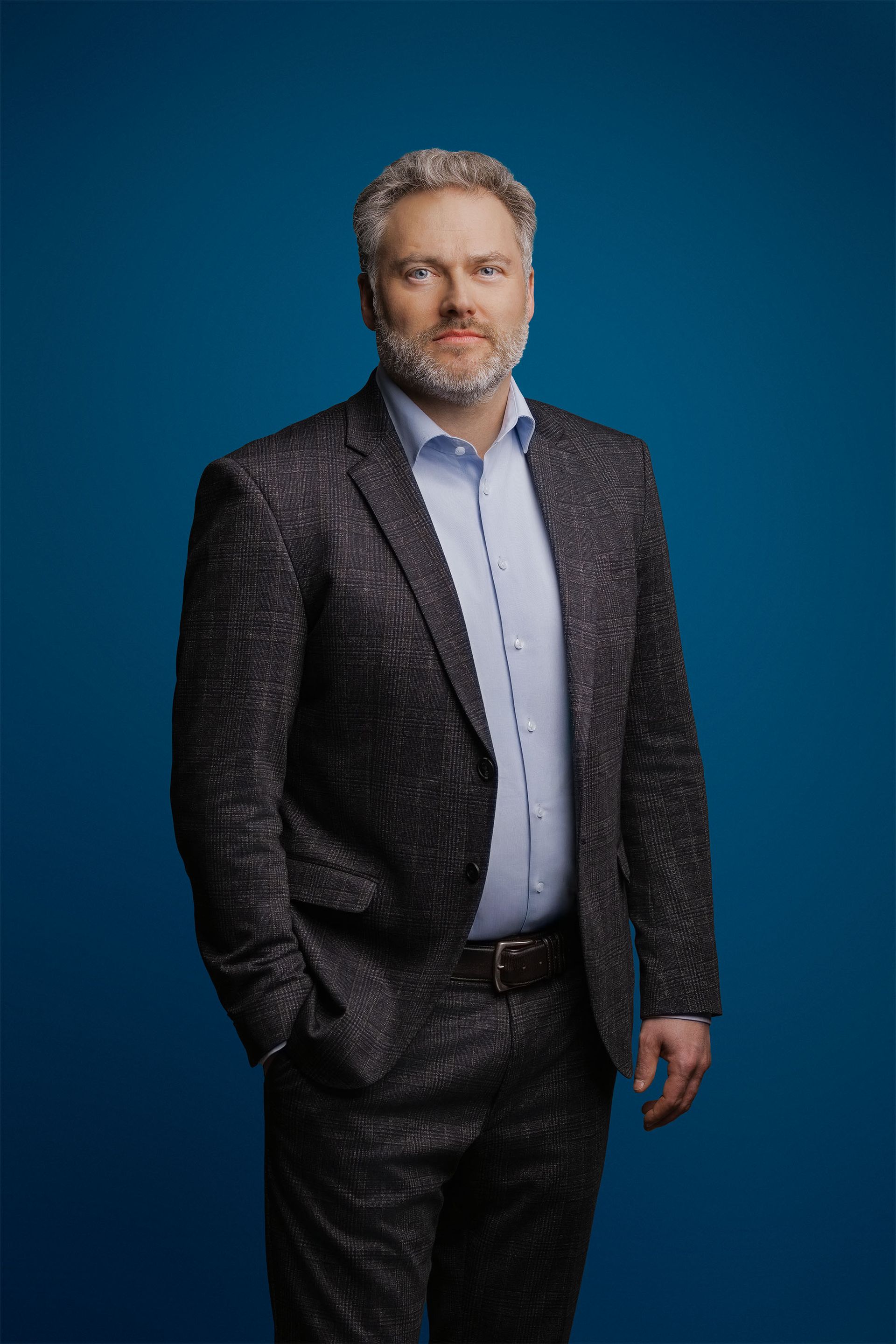 Eine Person mit Bart, die einen gemusterten Anzug und ein helles Hemd trägt, posiert vor einem einfarbig blauen Hintergrund.