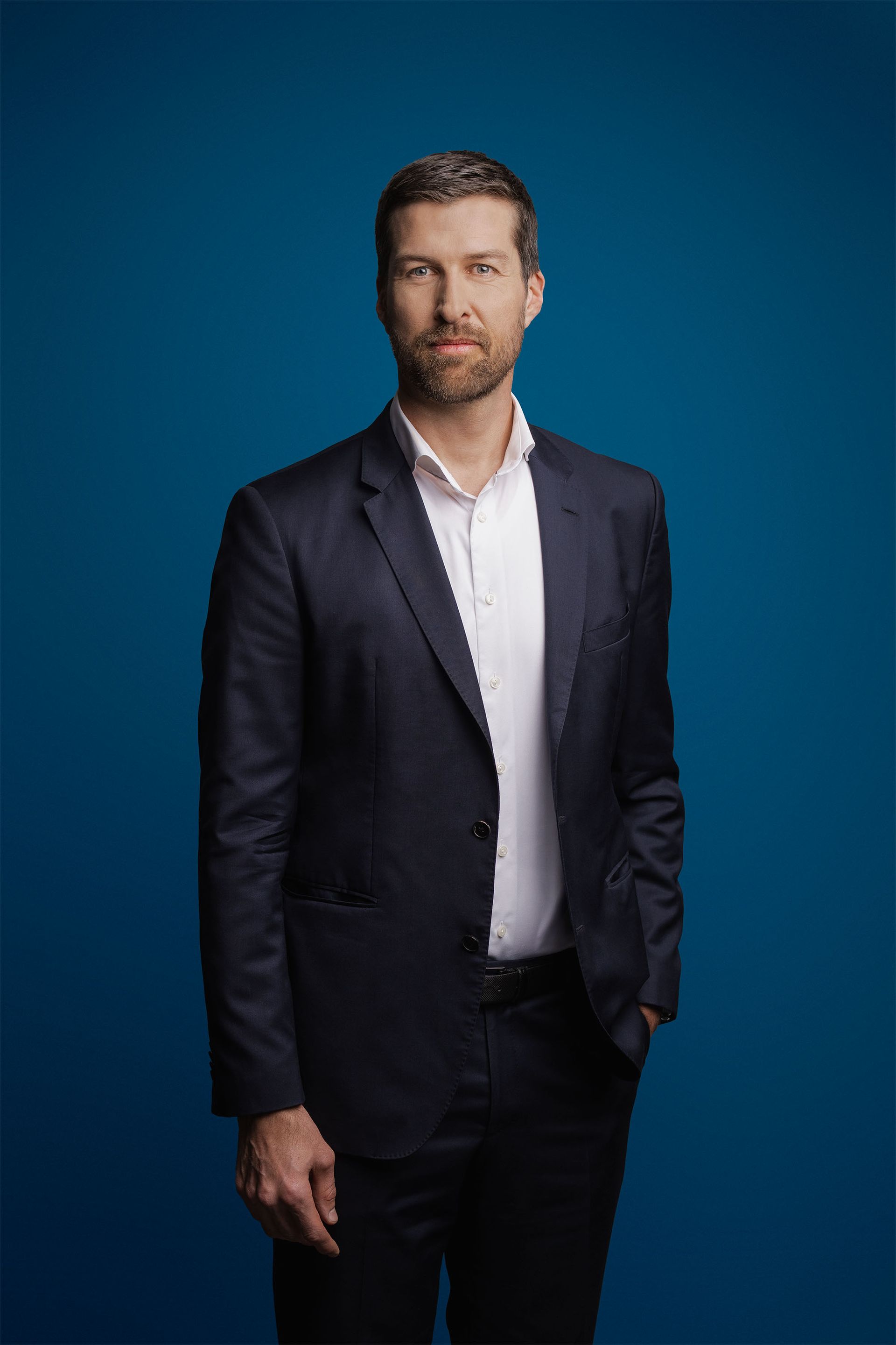 Ein Mann in einem dunklen Anzug und einem weißen Hemd steht vor einem einfarbig blauen Hintergrund und blickt in die Kamera.