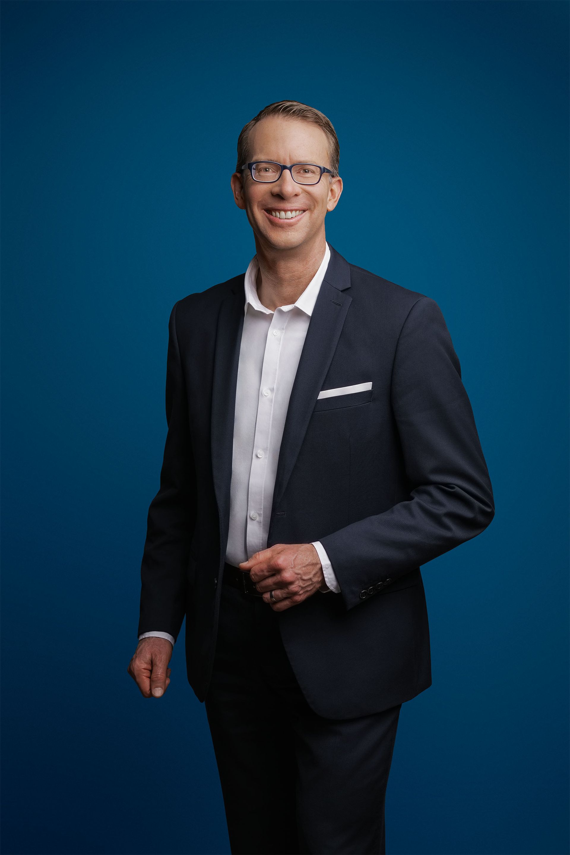 Lächelnde Person mit Brille und dunklem Anzug mit weißem Hemd vor einfarbig blauem Hintergrund.