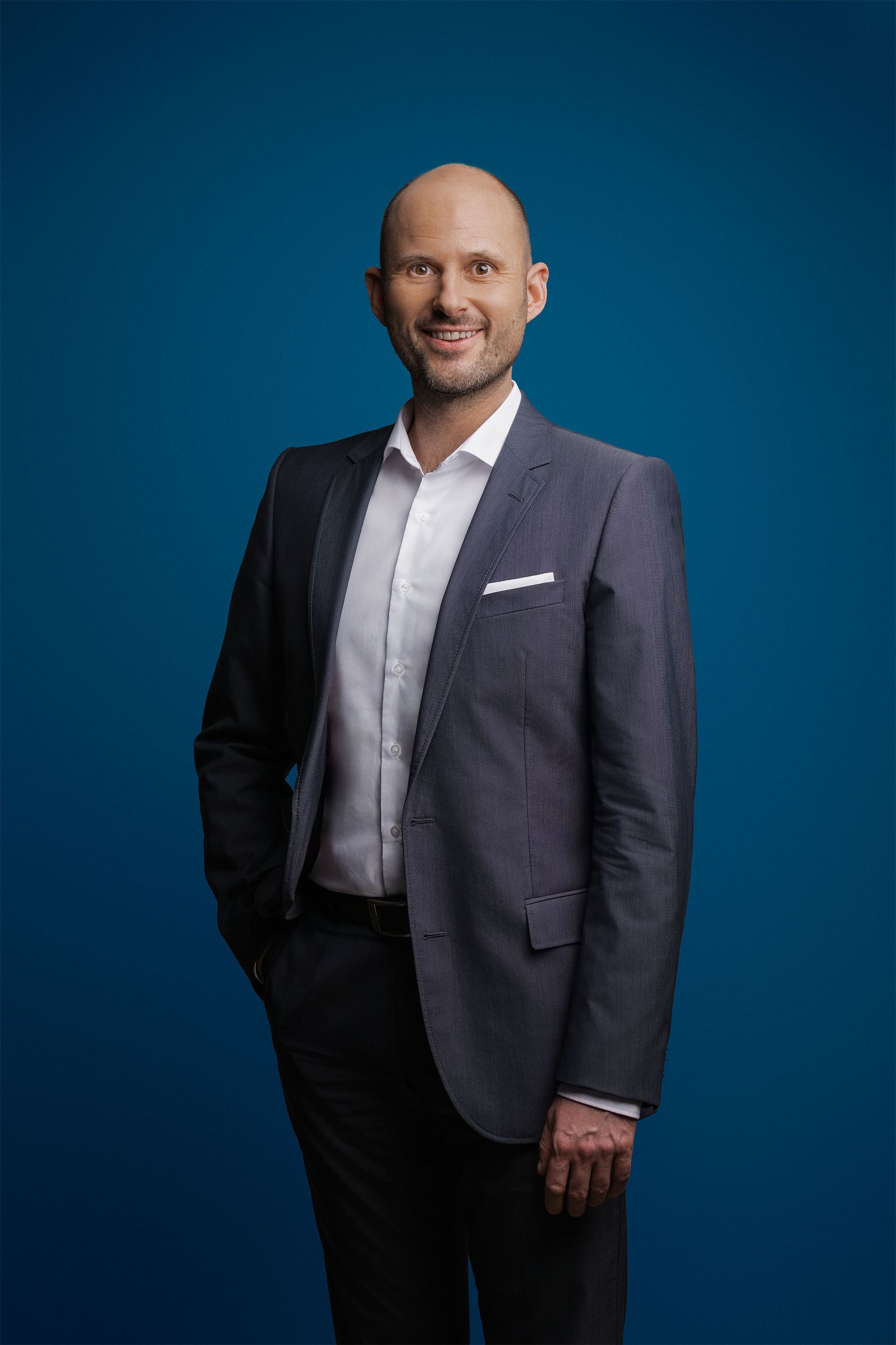 Eine lächelnde Person in einem blauen Blazer und einem weißen Hemd steht vor einem einfarbig blauen Hintergrund.