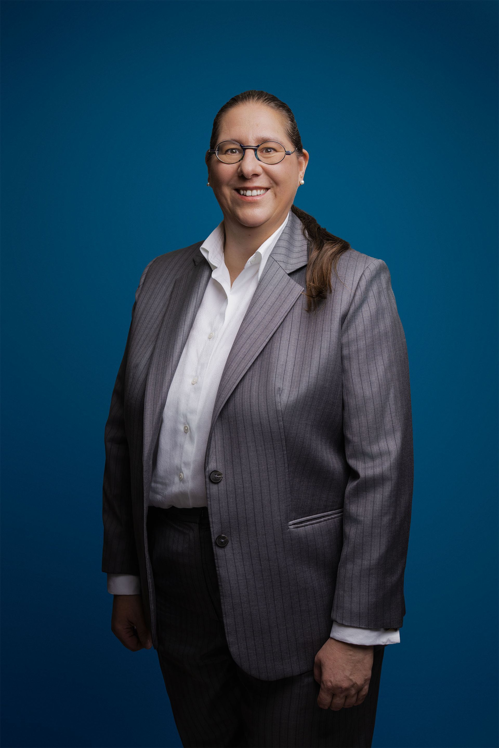 Eine Person mit langen dunklen Haaren und Brille trägt einen grauen Blazer und ein weißes Hemd vor einem schlichten blauen Hintergrund.