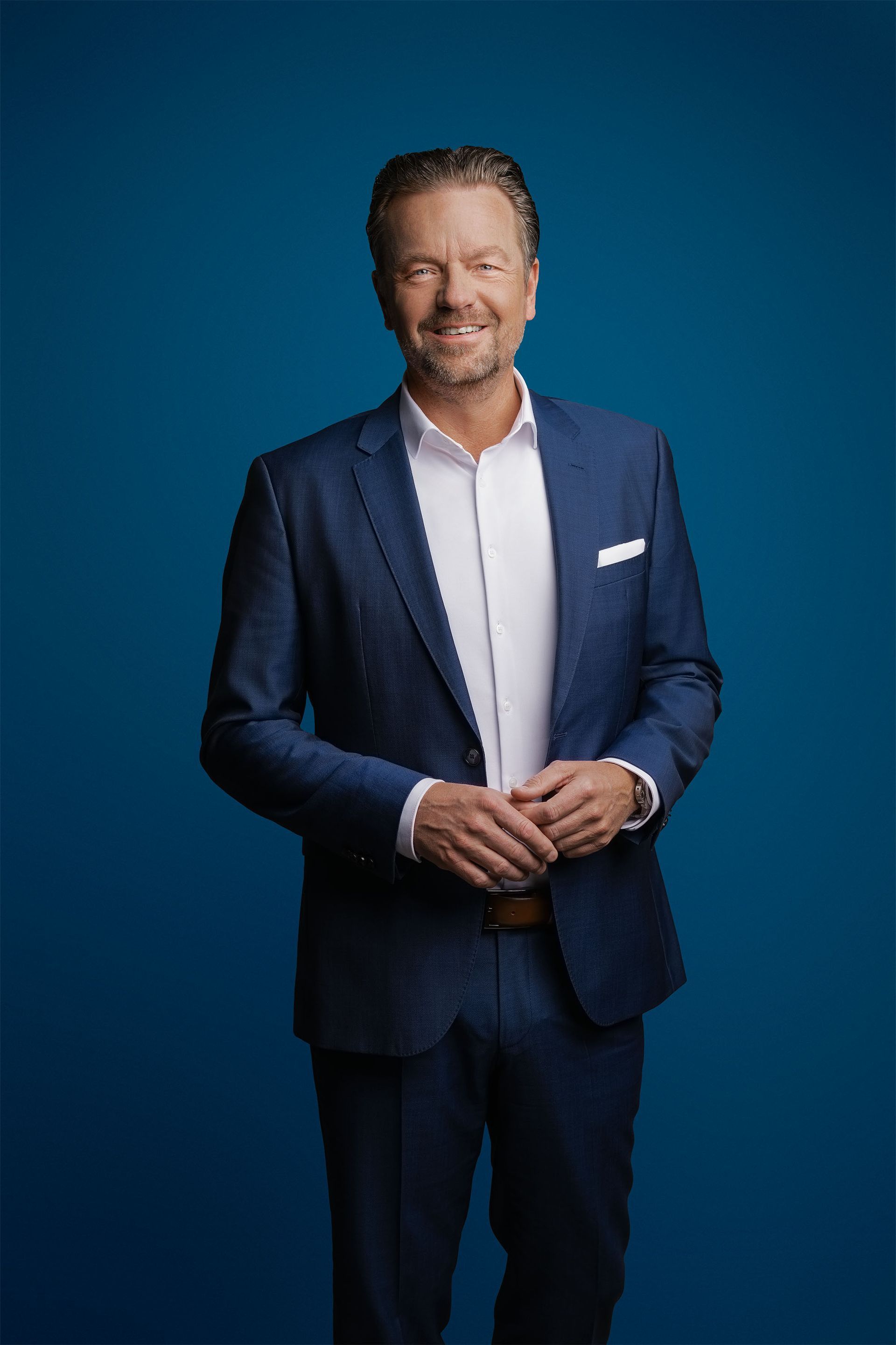 Eine lächelnde Person in einem dunkelblauen Anzug und weißem Hemd vor einem einfarbig blauen Hintergrund.