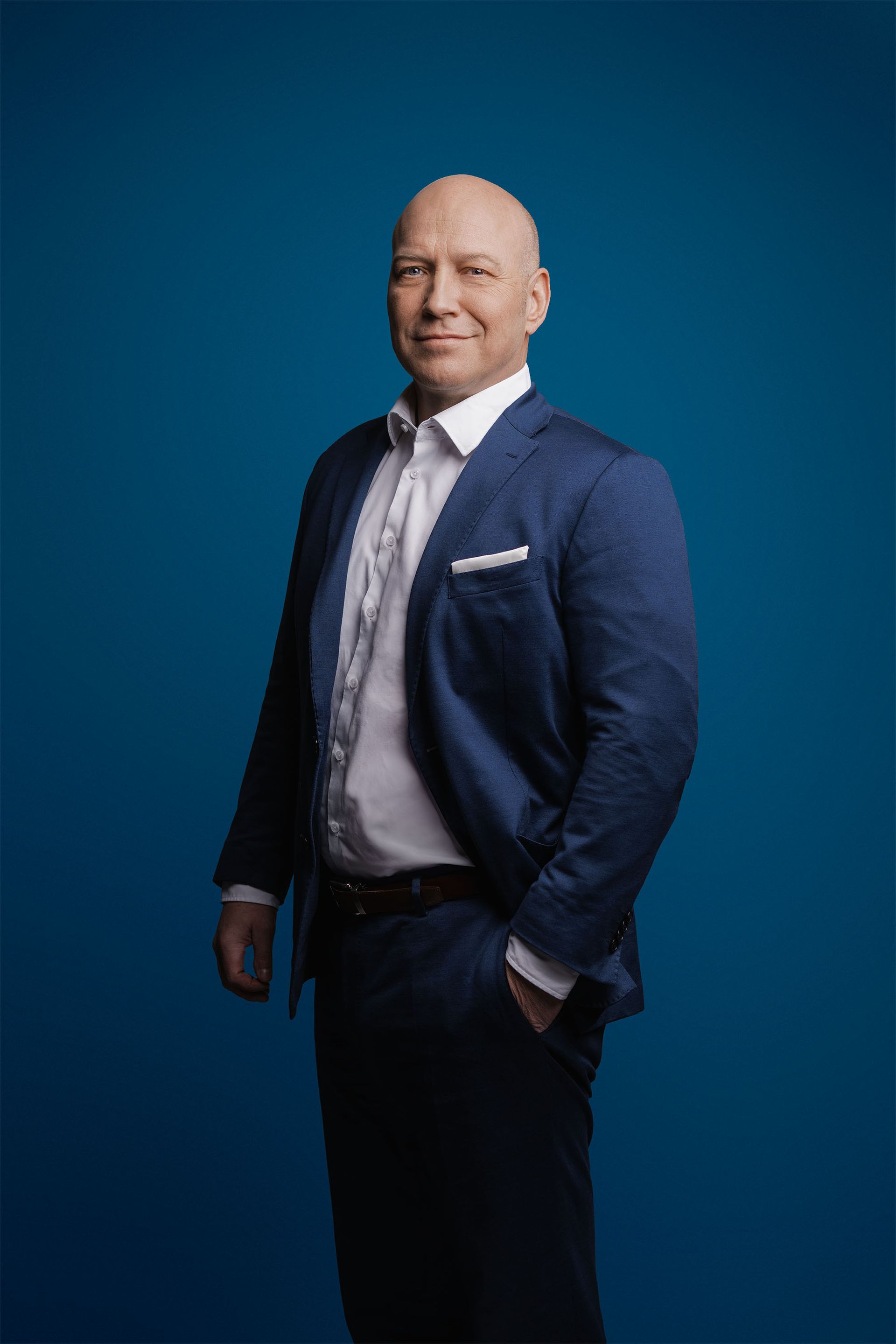 Ein glatzköpfiger Geschäftsmann in einem marineblauen Blazer und einem weißen Hemd steht vor einem einfarbig blauen Hintergrund.