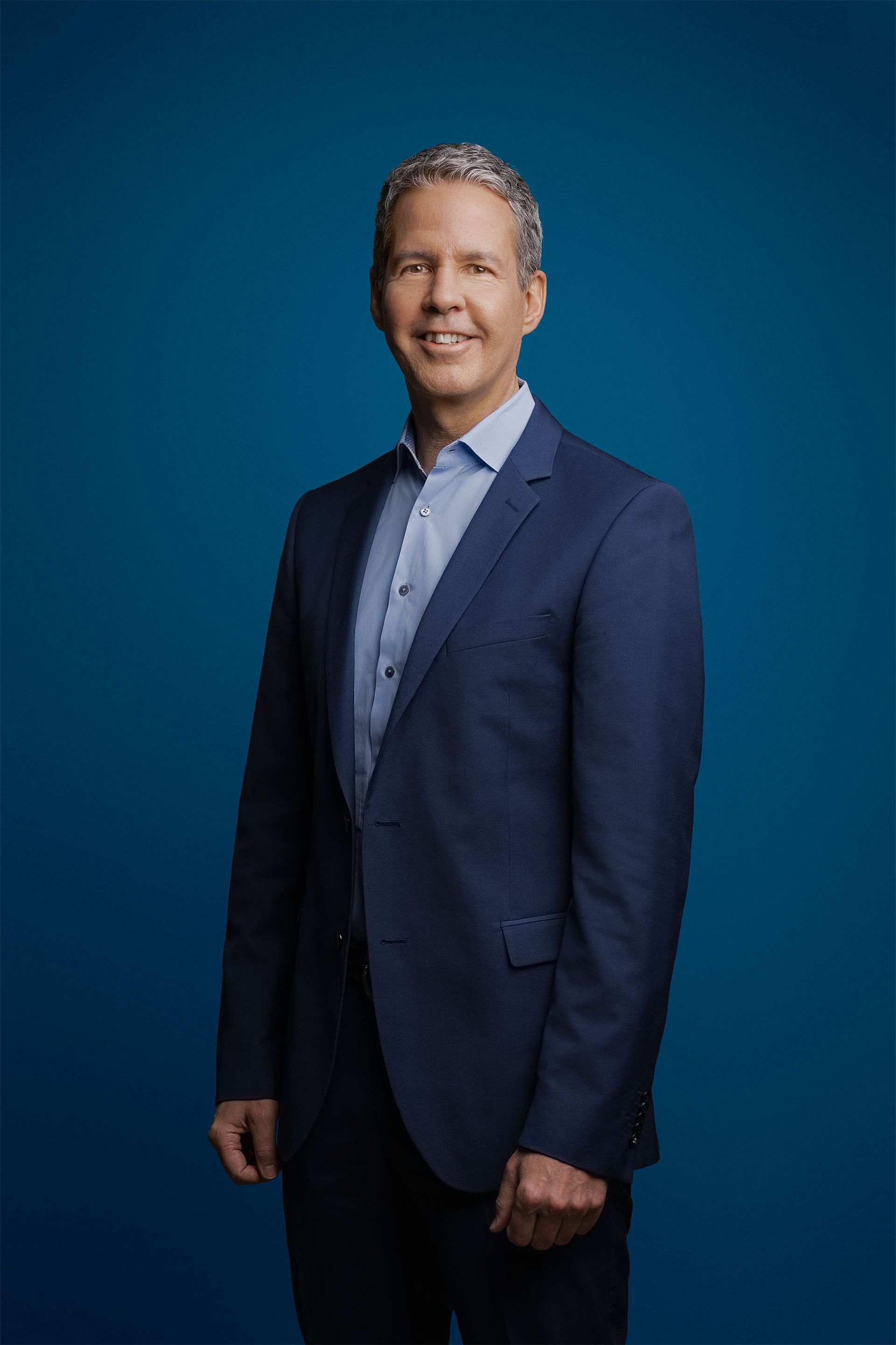 Eine Person in einem dunkelblauen Anzug und einem hellblauen Hemd mit Knöpfen steht vor einem einfarbig blauen Hintergrund und lächelt nach vorn.