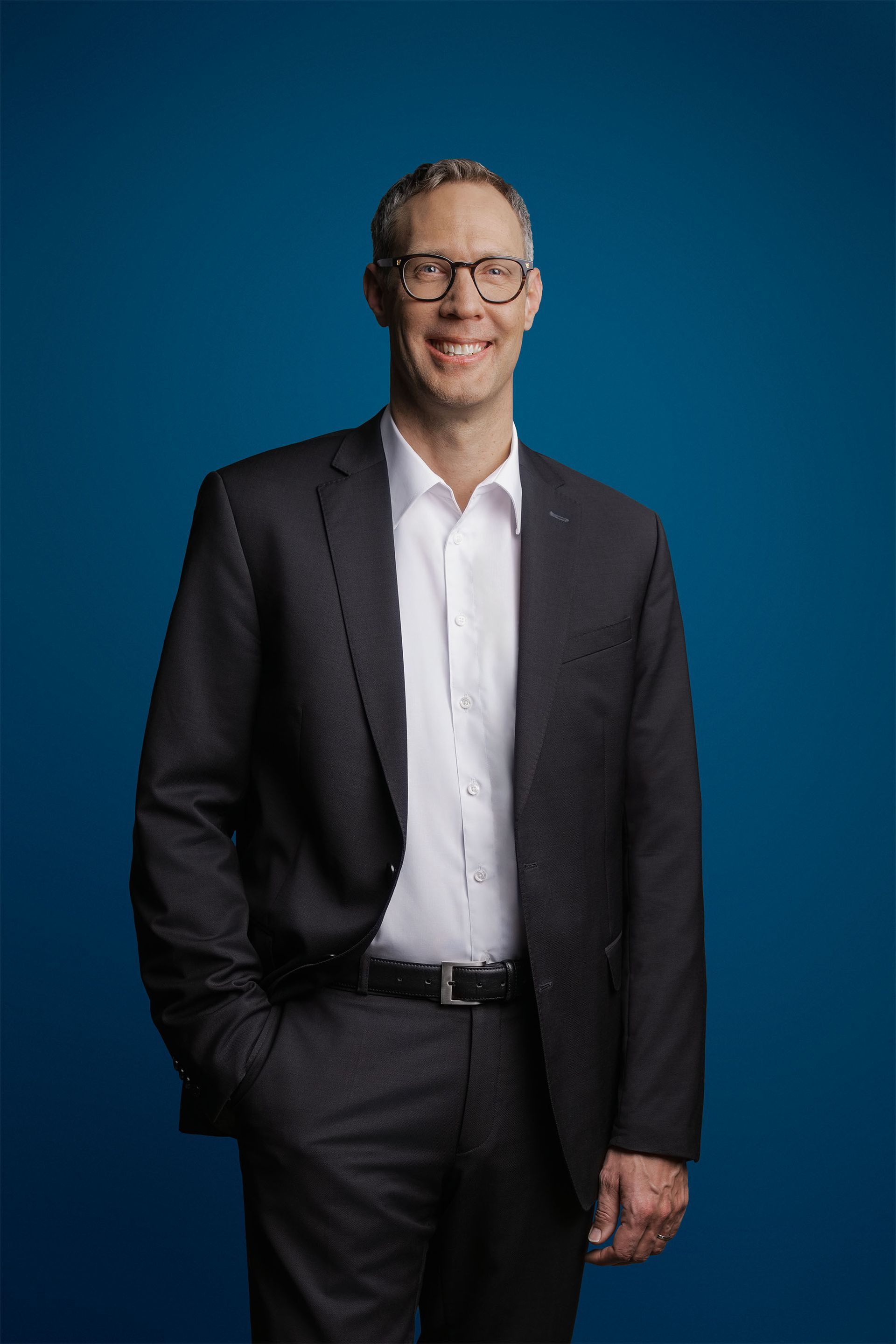 Eine Person in Anzug, weißem Hemd und Brille steht vor einem einfarbig blauen Hintergrund und lächelt in die Kamera.