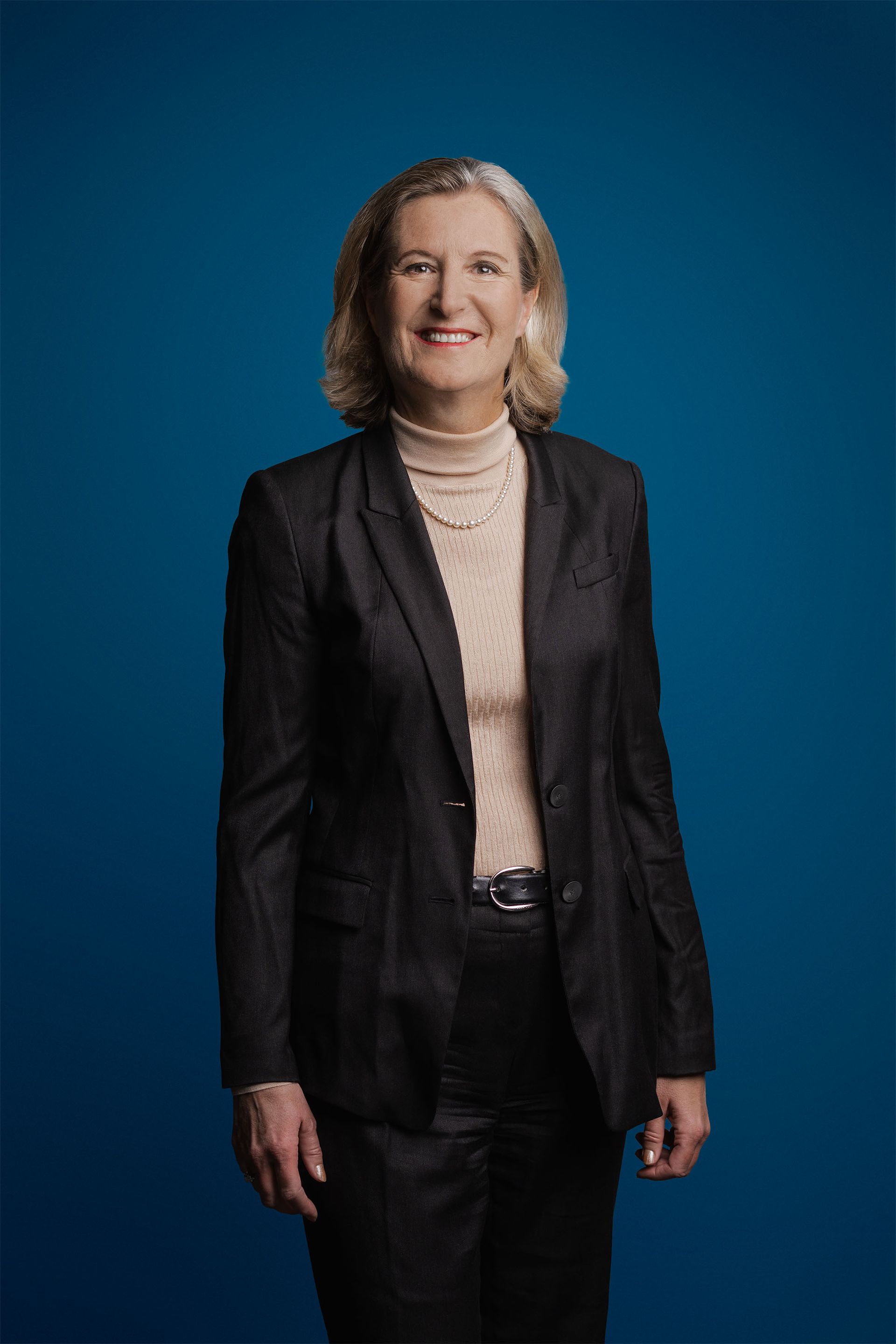 Ein professionelles Porträt einer Person in einem dunklen Blazer und einem neutralen Rollkragenpullover vor einem einfarbig blauen Hintergrund.