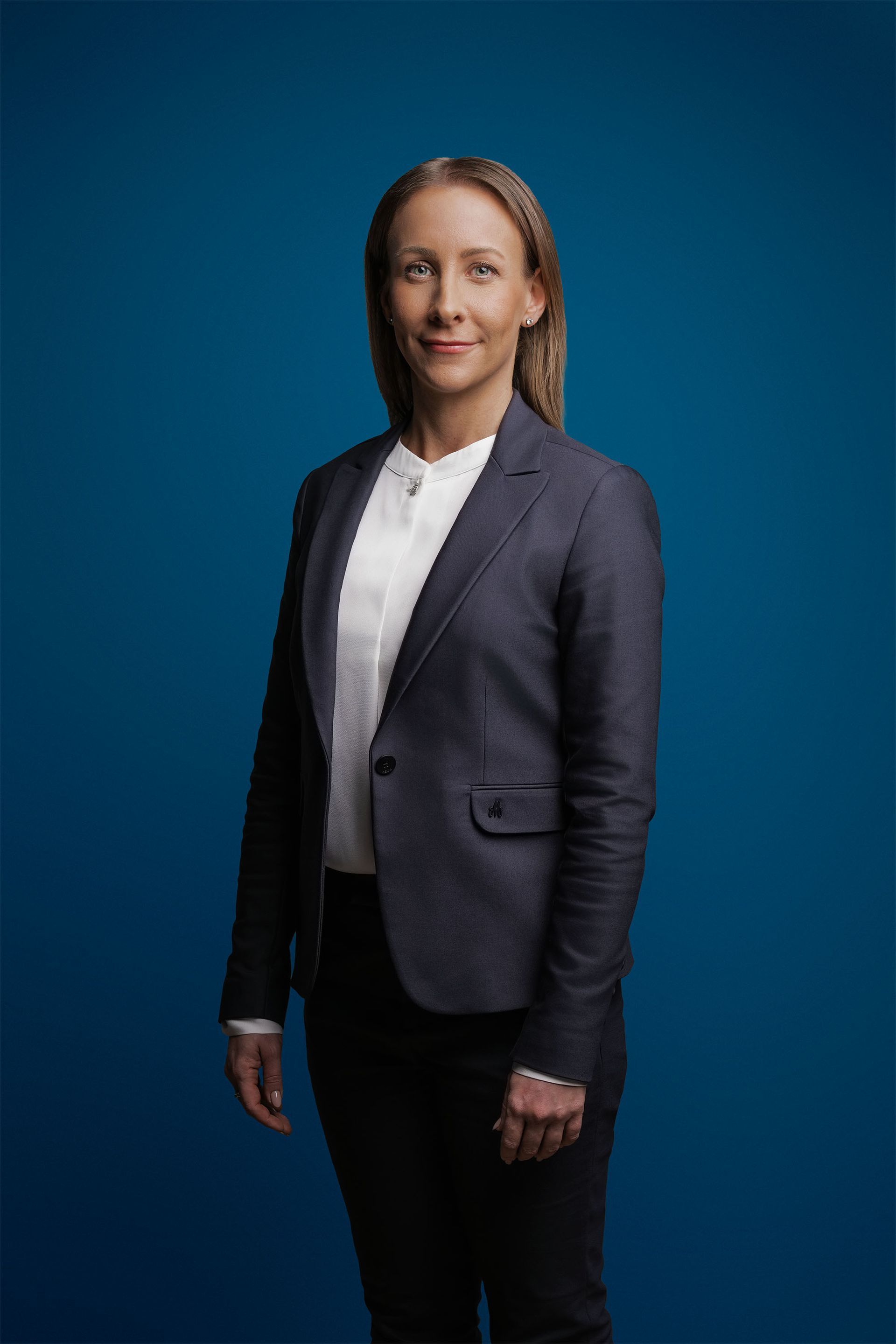 Eine Person in einem anthrazitfarbenen Blazer und weißem Hemd posiert vor einem einfarbig blauen Hintergrund und blickt in die Kamera.