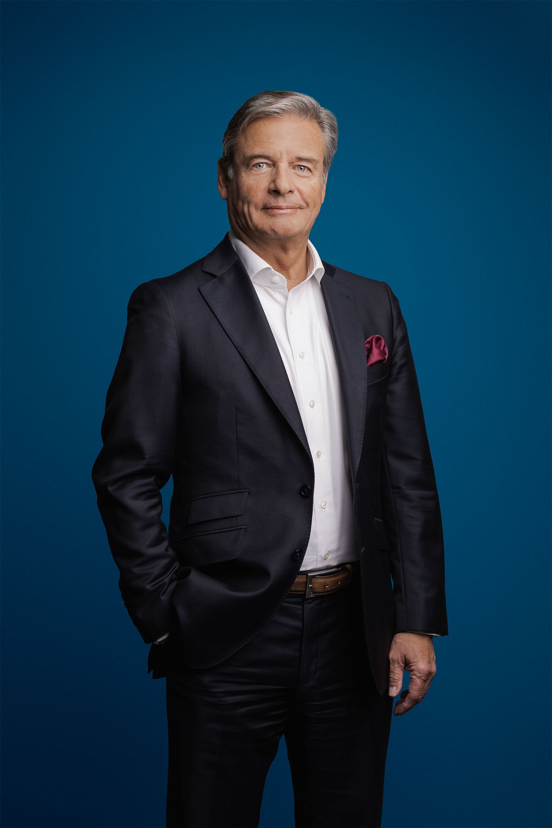 Ein professioneller Fotograf posiert vor blauem Hintergrund. Er trägt einen dunklen Blazer, ein weißes Hemd und ein rotes Einstecktuch.