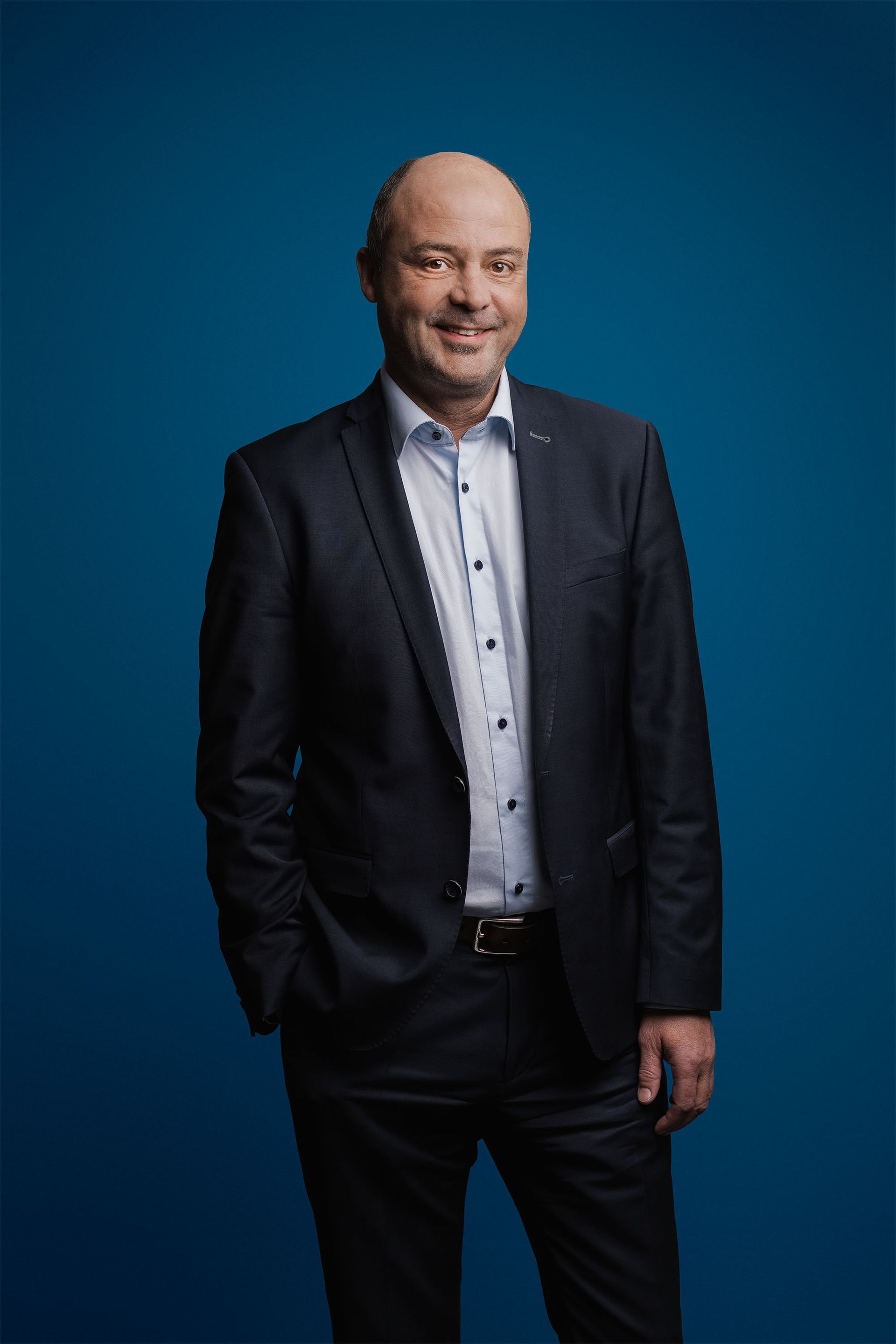 Ein Mann in einem dunklen Anzug und einem hellen Hemd mit Knöpfen lächelt vor einem einfarbig blauen Hintergrund.