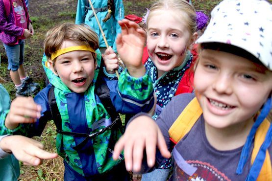 Drei Kinder mit fröhlichen Gesichtsausdrücken posieren im Freien, sie tragen Jacken und einen gemusterten Sonnenhut, während sie in einem Wald wandern.