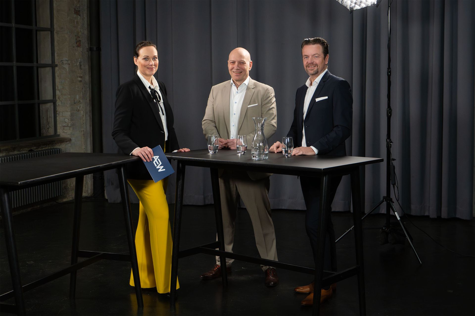 Drei Personen stehen hinter einem hohen schwarzen Tisch in einem Studio mit grauen Vorhängen und lächeln in die Kamera.