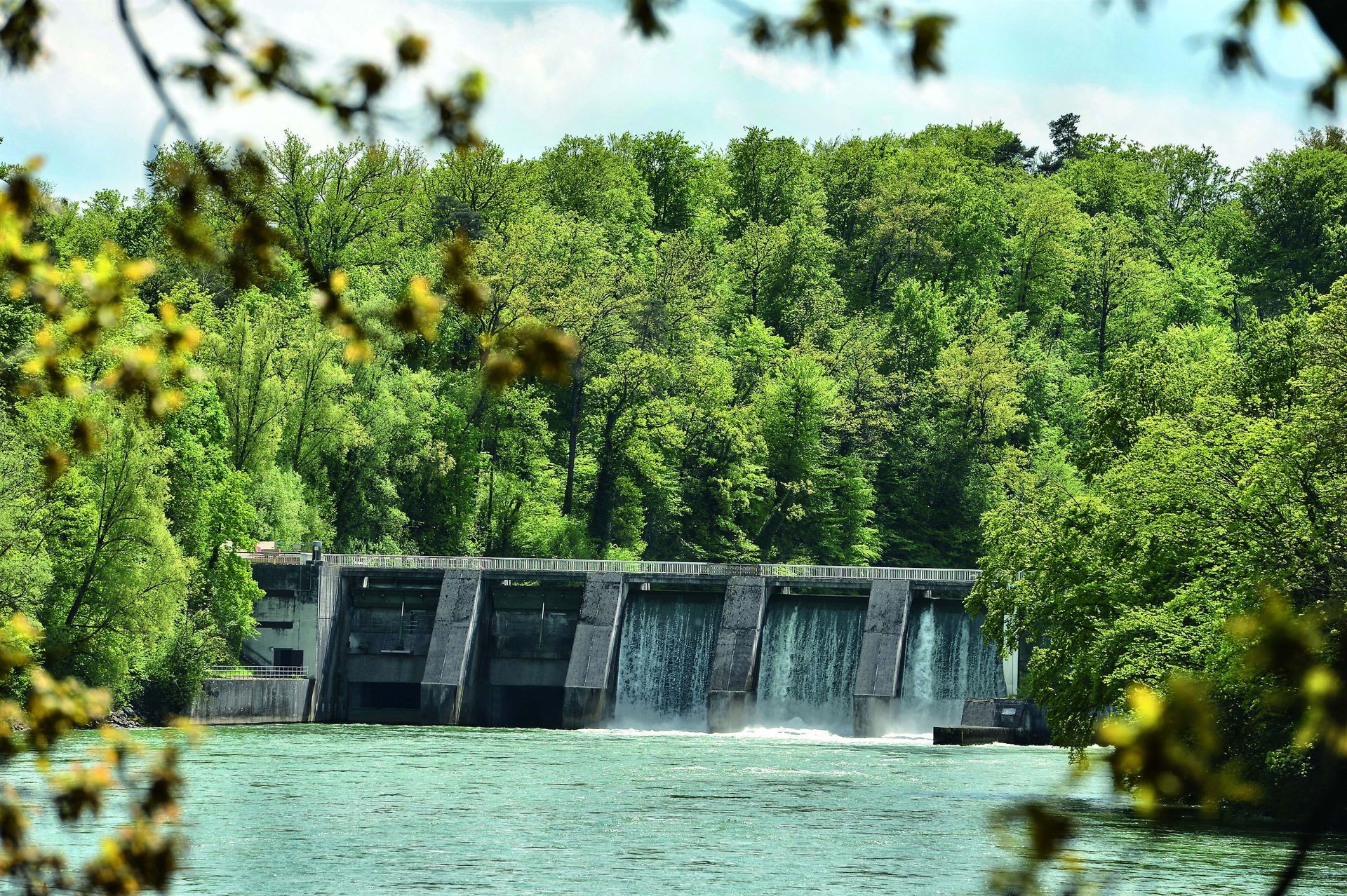 Ein Betonwehr überspannt einen Fluss, über dessen Abschnitte das Wasser kaskadenartig herabstürzt, vor dem Hintergrund eines dichten, üppig grünen Baumbestands.