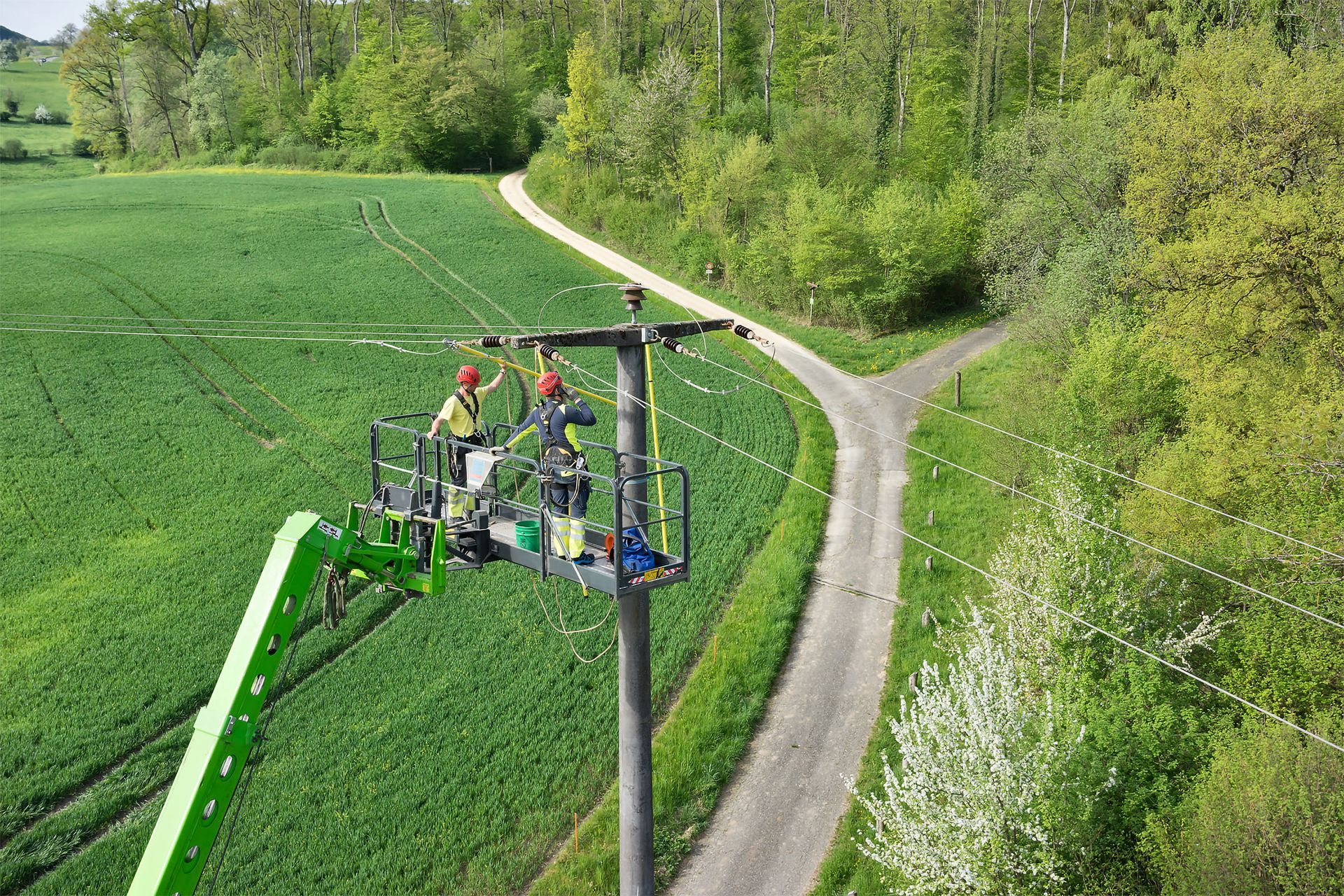 Zwei Arbeiter eines Versorgungsunternehmens führen in einem grünen Hubsteiger Wartungsarbeiten an einem Strommast durch, der einen Feld- und Waldweg überblickt.