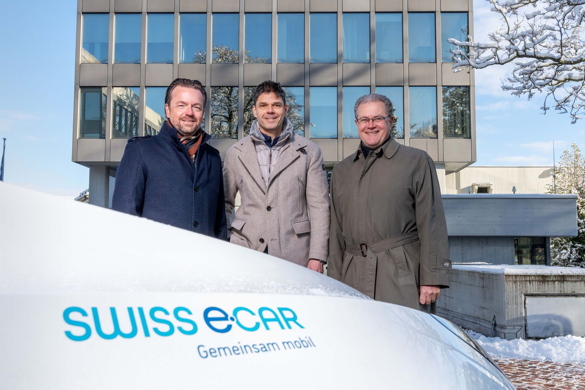 Drei Männer stehen an einem verschneiten Tag vor einem Gebäude neben einem weißen Fahrzeug mit dem Logo „SWISS e-CAR“.