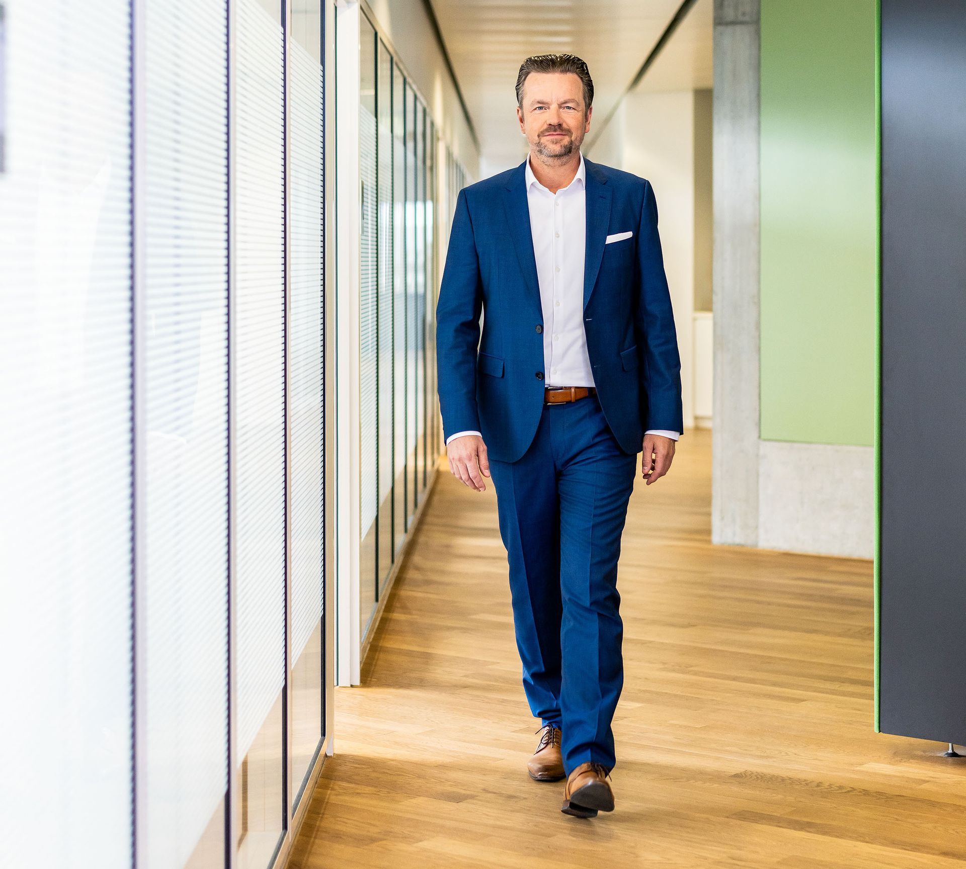 Eine Person in einem blauen Anzug und weißem Hemd geht einen modernen Büroflur mit Holzboden und Glaswänden entlang.