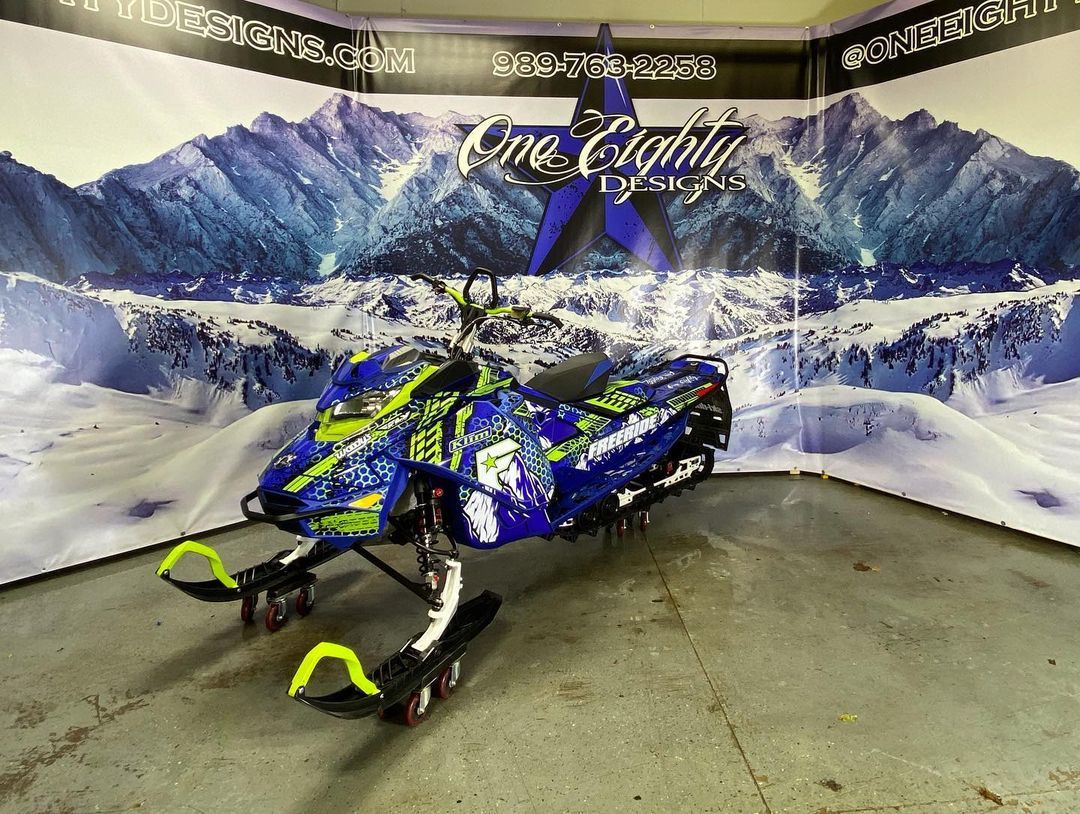 A blue and green snowmobile is parked in front of a wall with mountains in the background.