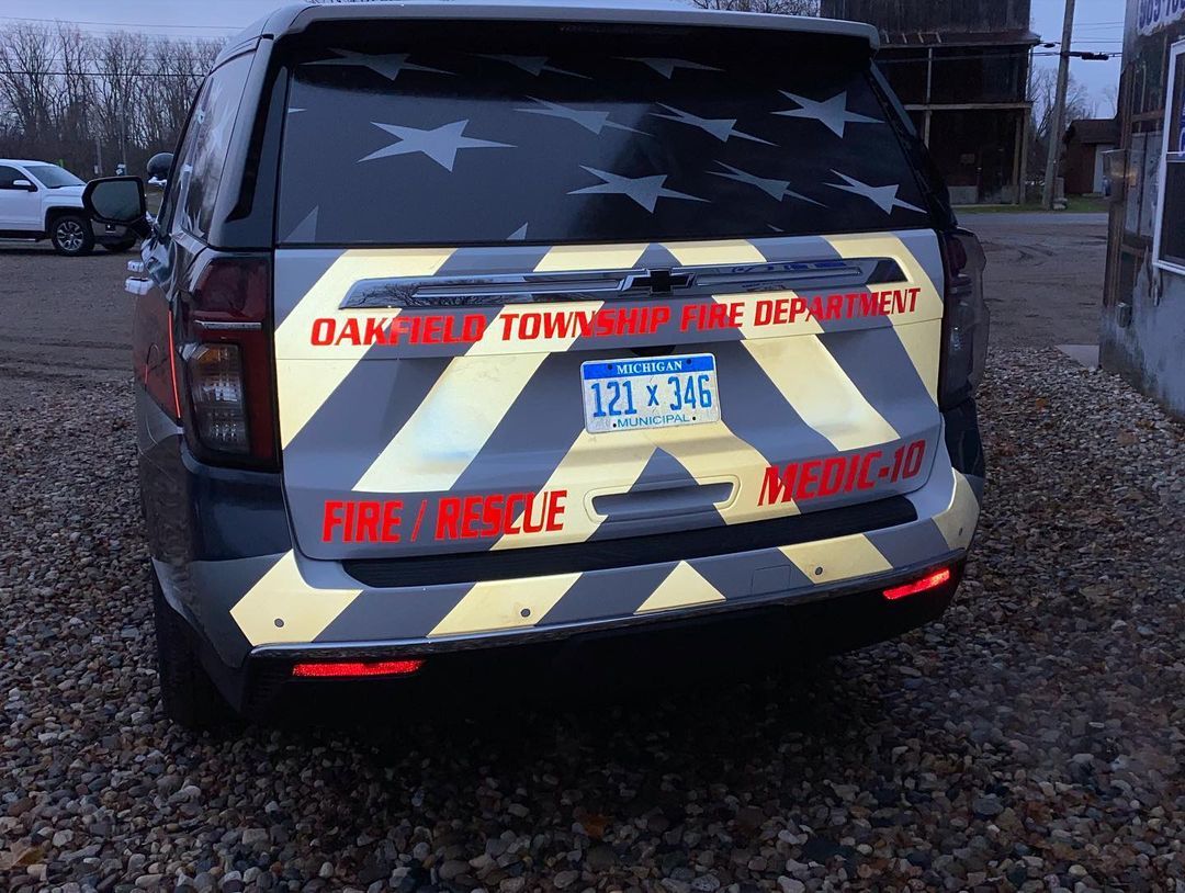 A gray and yellow suv with a flag on the back is parked in a gravel lot.