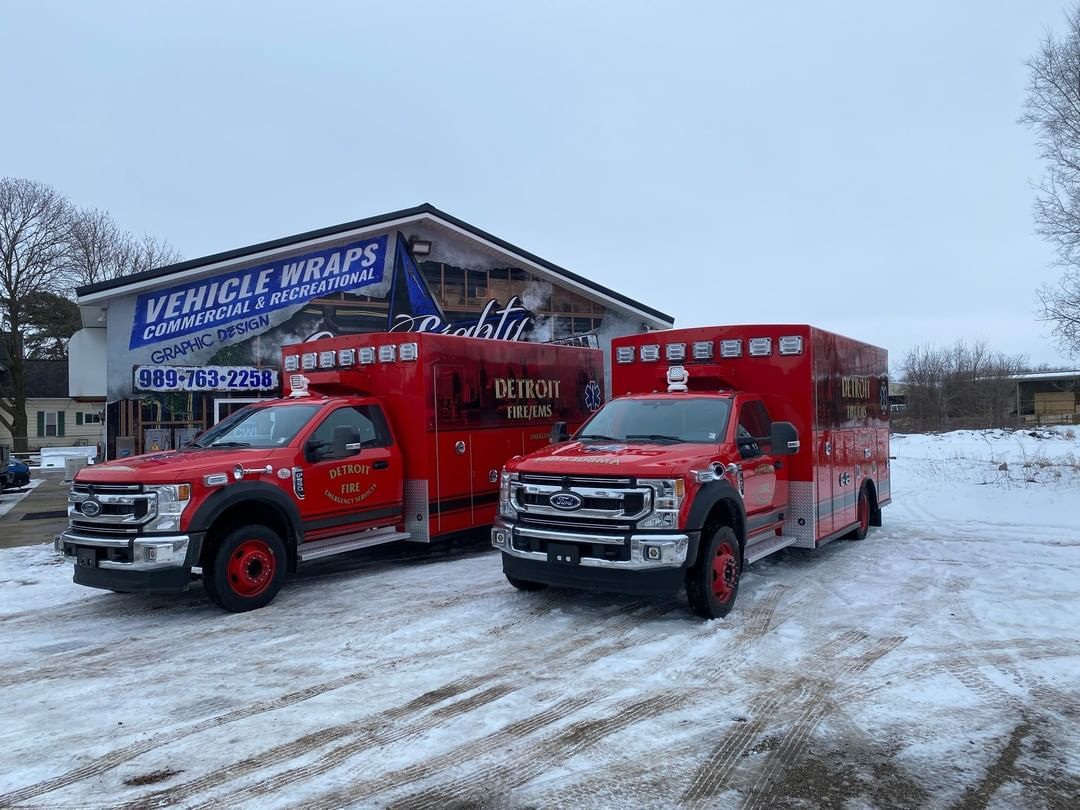 Two red ambulance trucks are parked in the snow in front of a building.