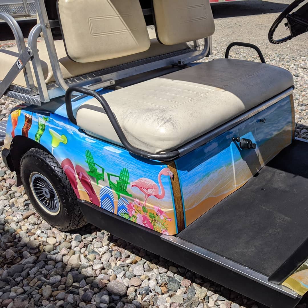 A golf cart with flamingos painted on the side is parked on gravel.