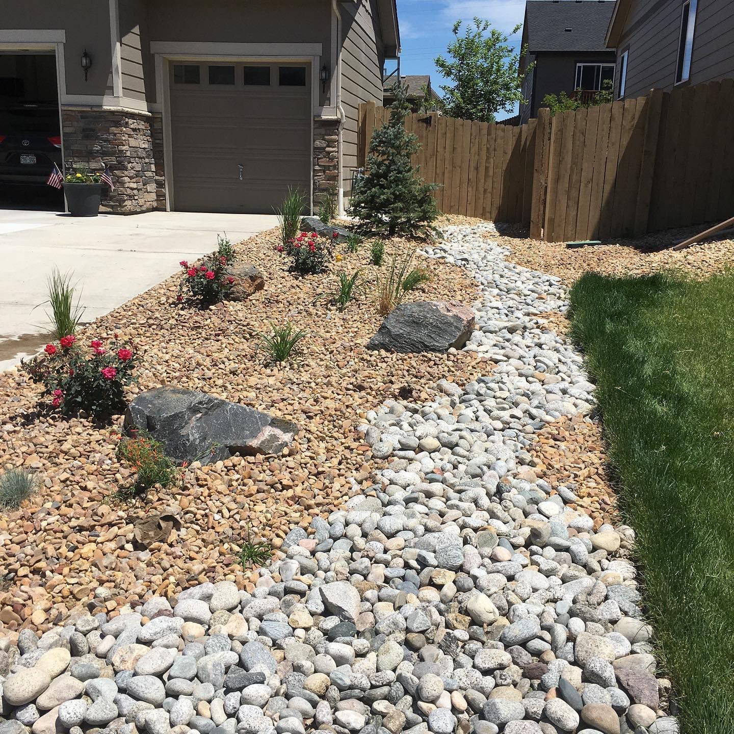 Front yard landscaping with a rock-filled dry creek bed, surrounded by brown gravel, plants, and a wooden fence.