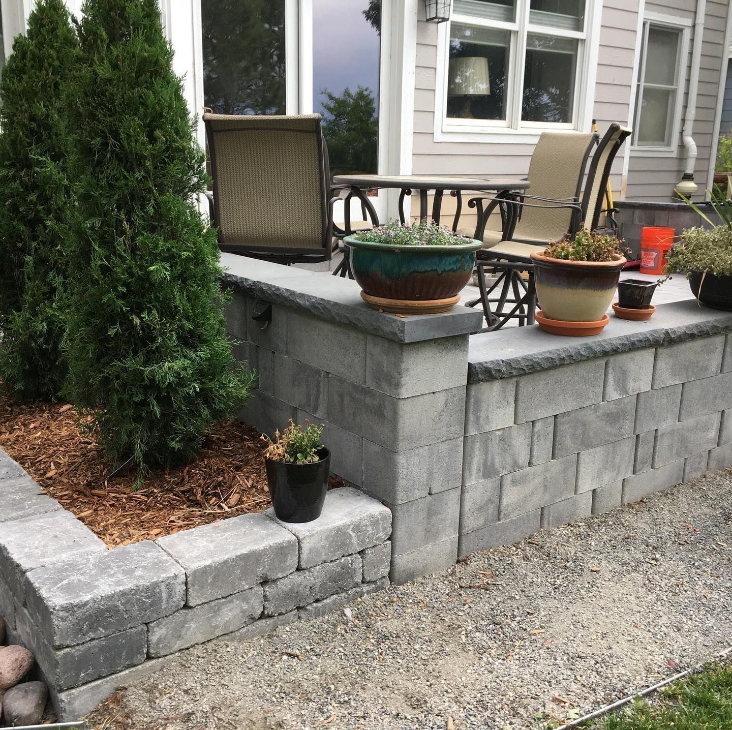 A patio area with a retaining wall made of gray blocks. Two green evergreen trees are planted in the bed.