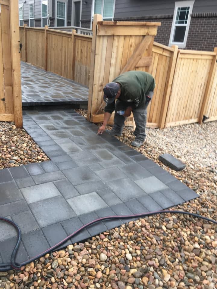 Man laying paving stones in a yard, near a wooden fence and gate. Stones are dark grey and brown gravel surrounds them.