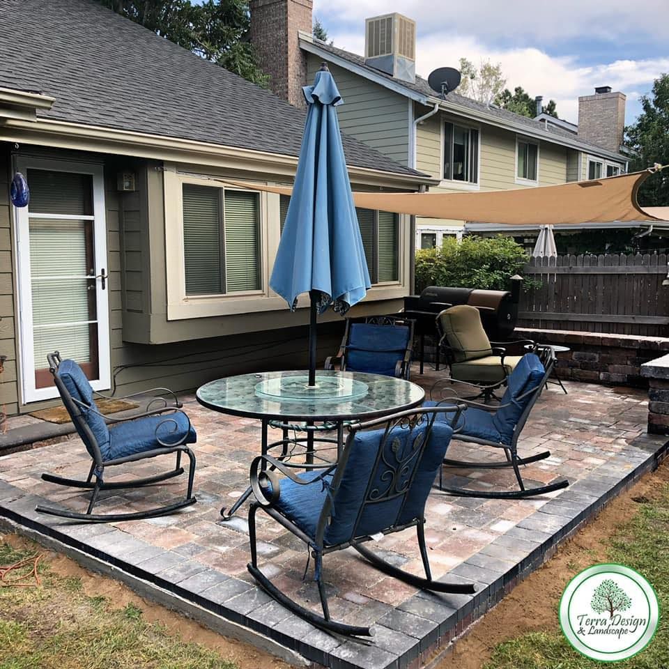 Patio with a glass table, blue rocking chairs, and a large umbrella. There's a sunshade over more seating and a grassy yard.