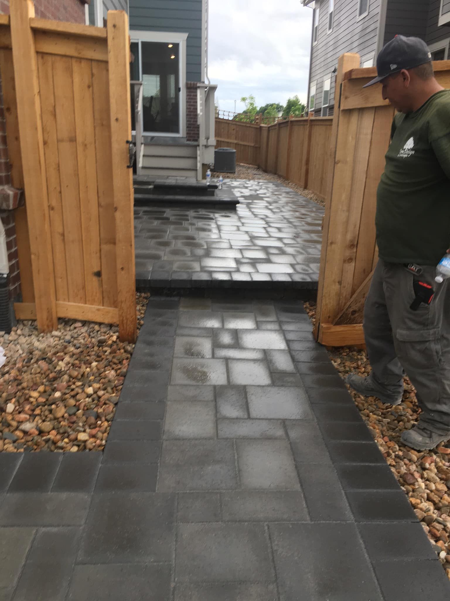 A paved walkway leading to a house, framed by wooden fences and landscaping. A man stands to the right, looking down.