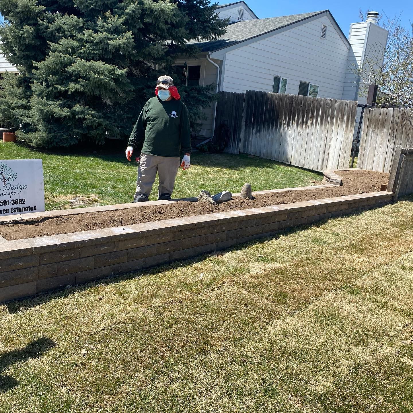 A person wearing a mask stands in a sunny backyard garden bed filled with soil. They are wearing a green shirt and khakis.