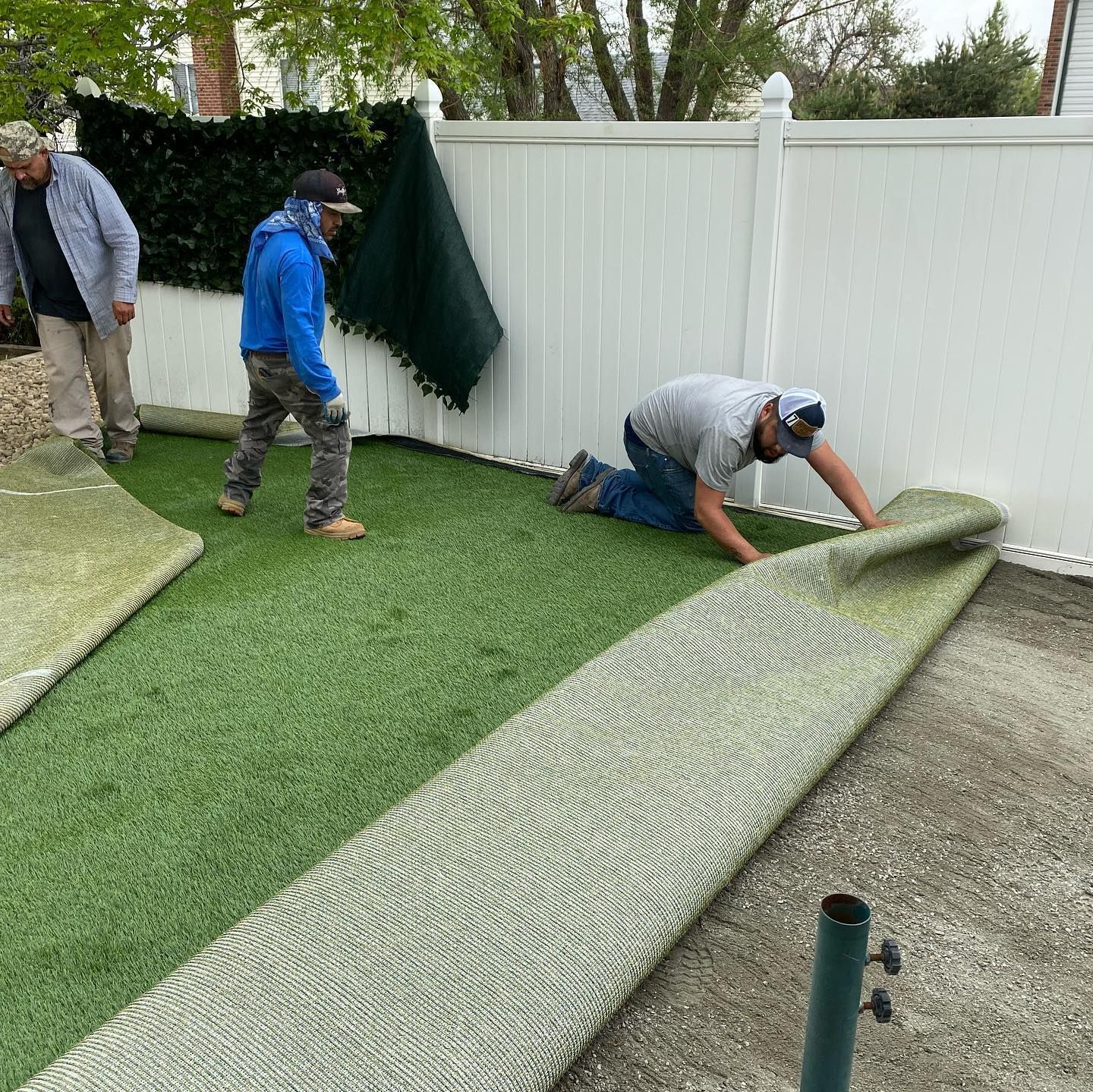 Three workers installing artificial turf against a white fence. One unrolls the turf, while the others stand nearby.