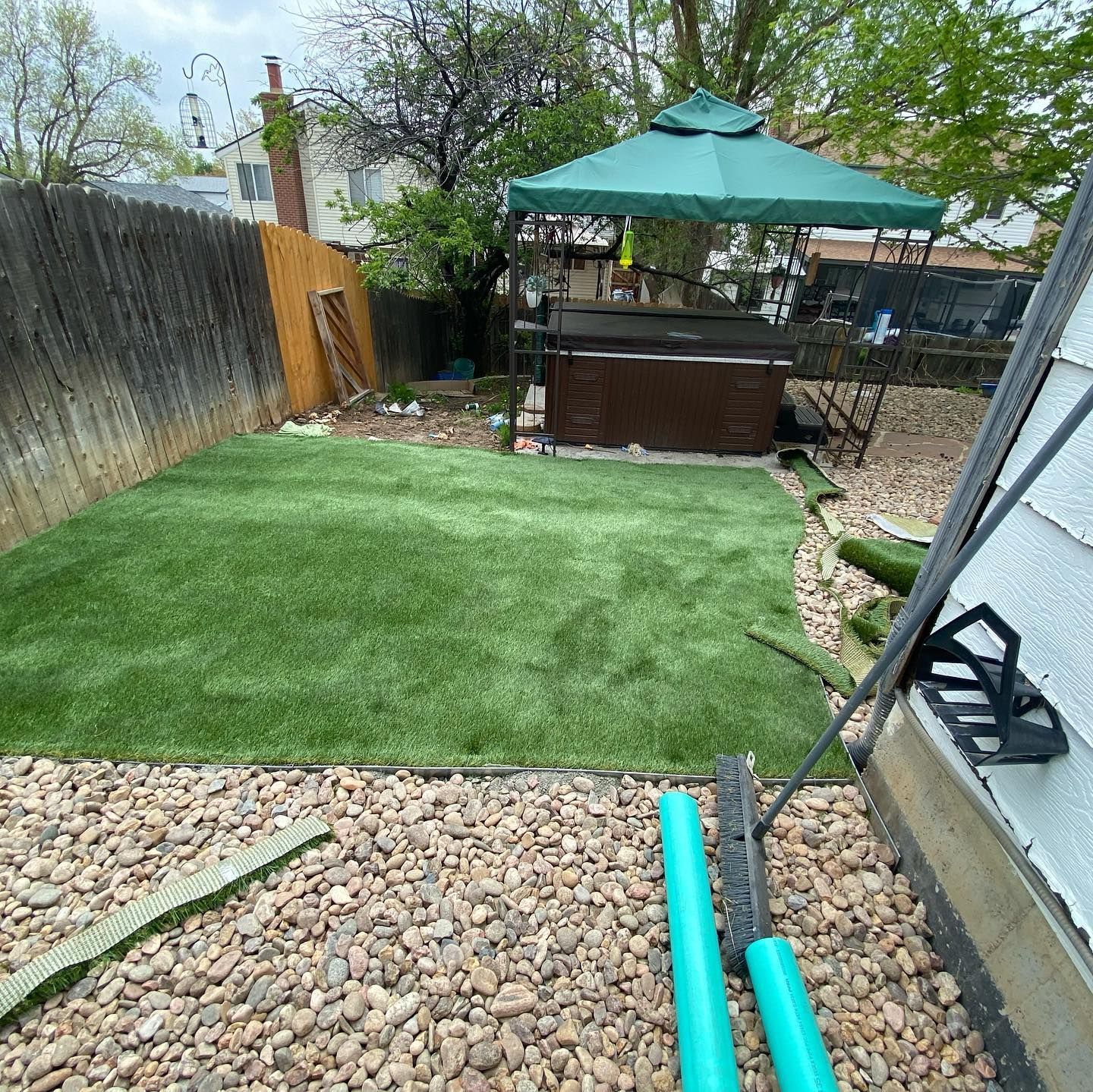 Backyard with a patch of green artificial turf, a hot tub under a green umbrella, and a rock border.