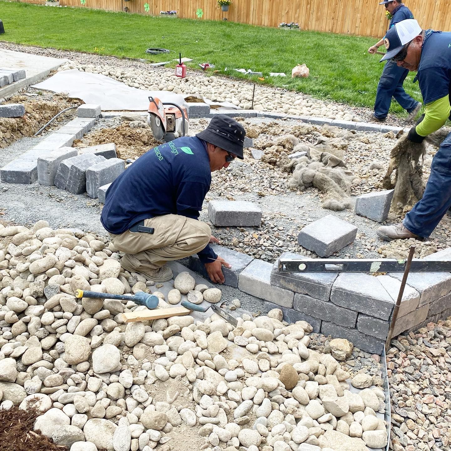 Construction workers laying paving stones outdoors, surrounded by gravel and tools. One worker kneels, placing a stone.