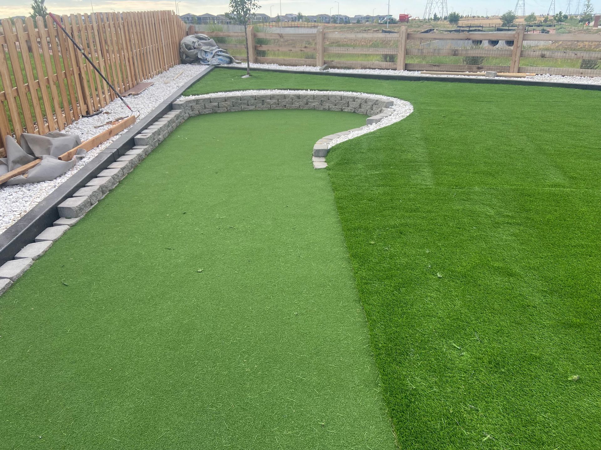 A putting green with artificial turf and a stone border in a backyard, next to a wooden fence.