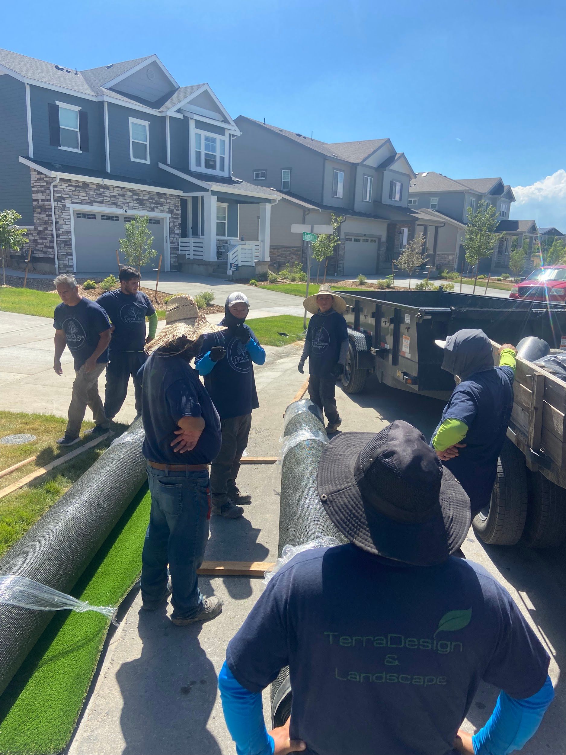 A group of workers unloads rolls of artificial turf from a truck in front of houses.