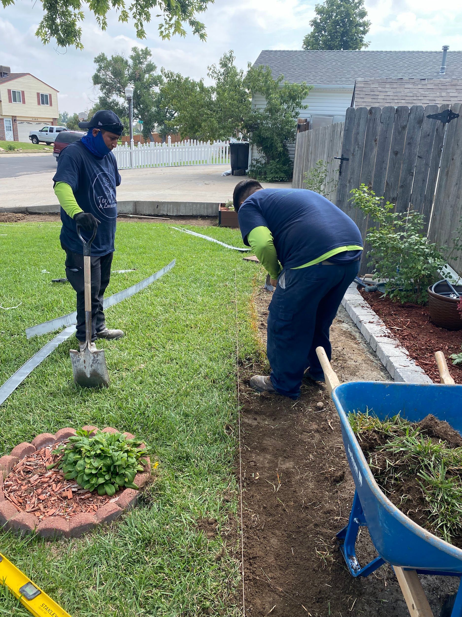 Two workers install landscaping edging in a residential yard. One digs with a shovel, while the other appears to measure.
