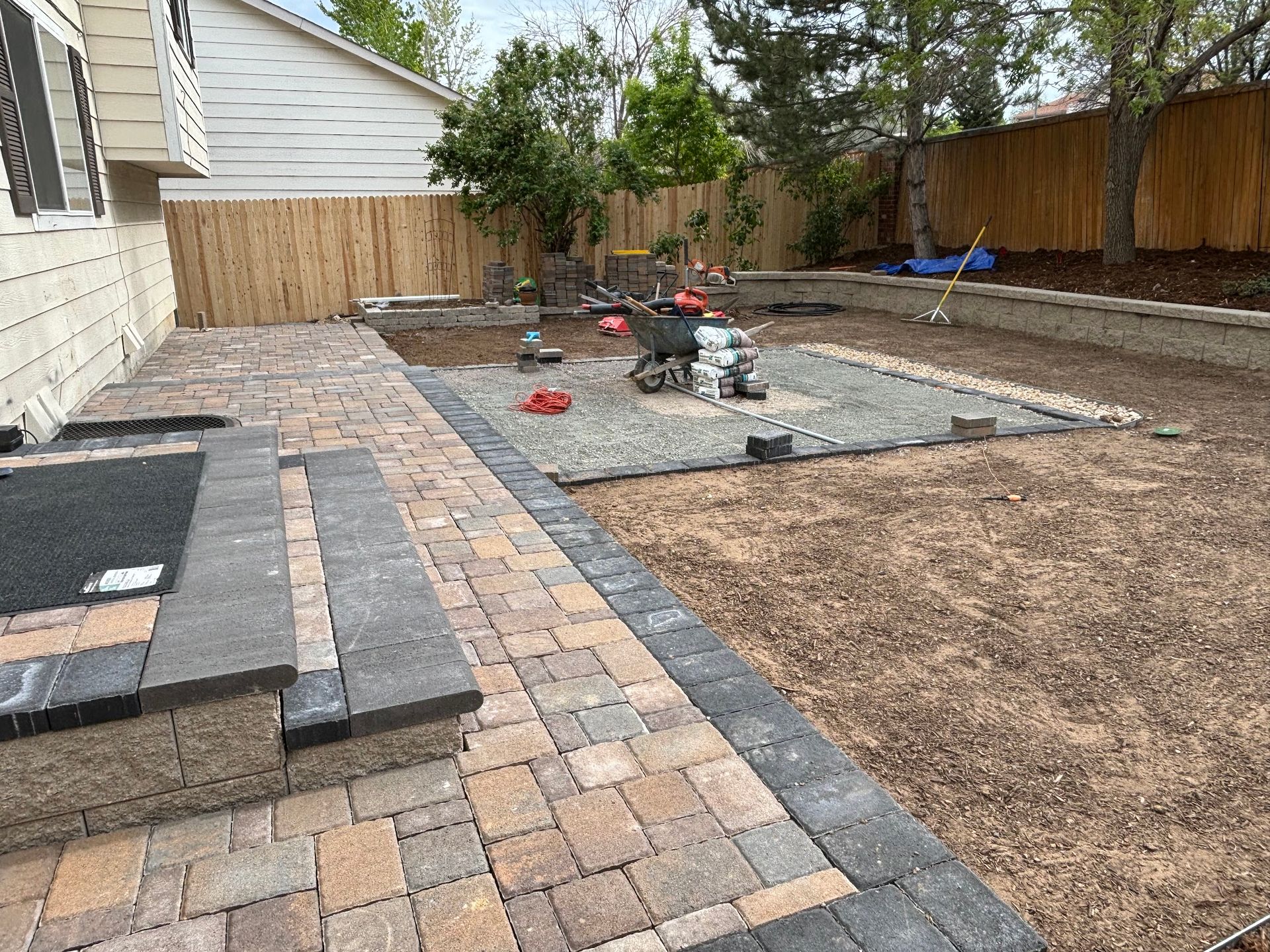 A backyard under construction with a new stone patio, retaining wall, and gravel area.