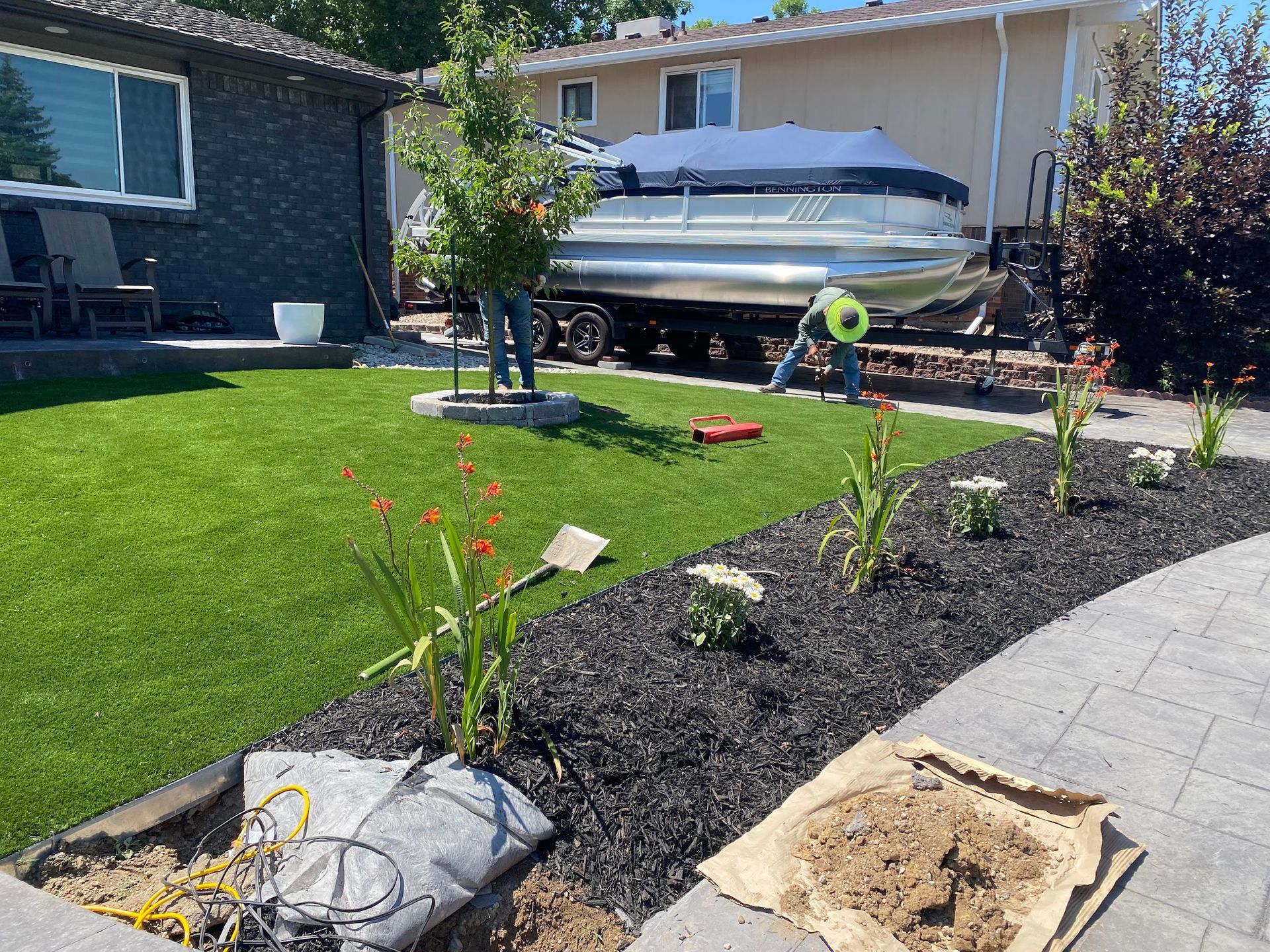 A person works on landscaping near a boat on a trailer. Black mulch and flowers border the green lawn and paved walkway.