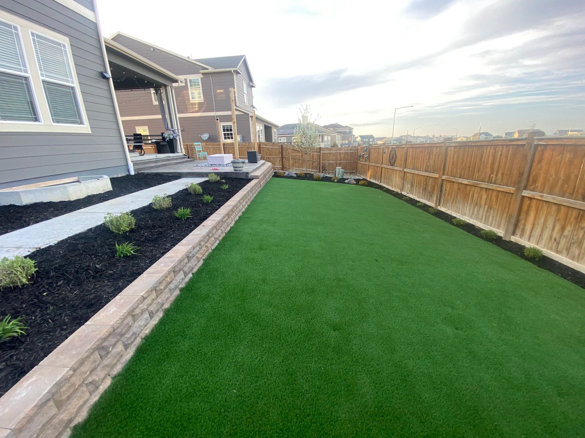 Backyard with artificial green turf, stone retaining wall, wooden fence, and two-story houses under a cloudy sky.