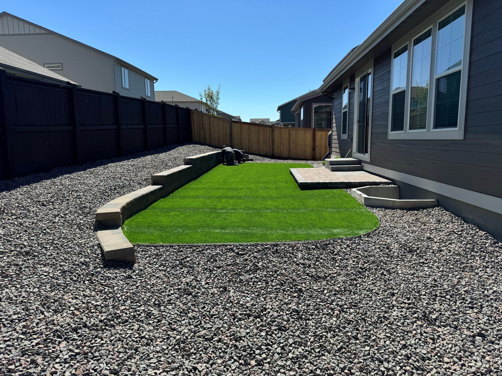 Backyard with a small patch of green turf surrounded by dark gray gravel, concrete retaining walls, and a wooden fence.