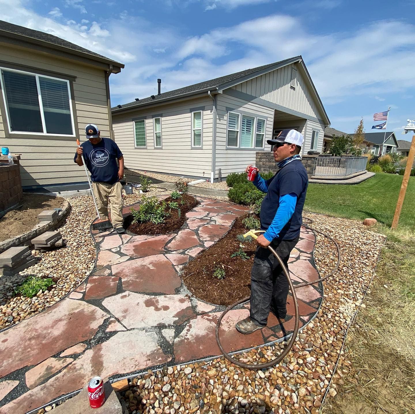 Two men landscaping a garden bed with flagstone path, plants, and mulch. One waters plants with a hose.