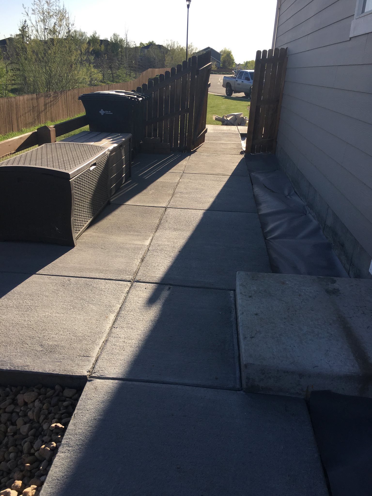 A gray paved walkway next to a light gray house leads toward a street. A dark fence and a garbage bin are on the left.